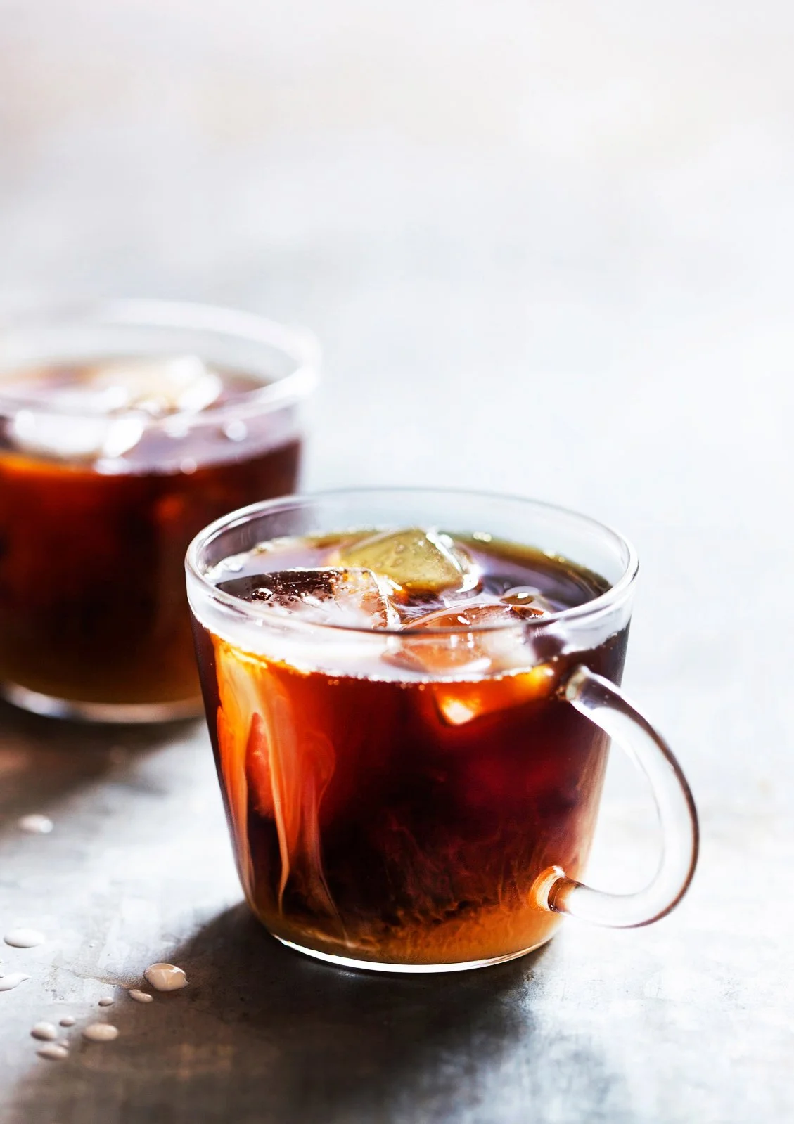 Two clear glass cups filled with iced coffee or tea, sitting on a light surface, with ice cubes visible and scattered around.