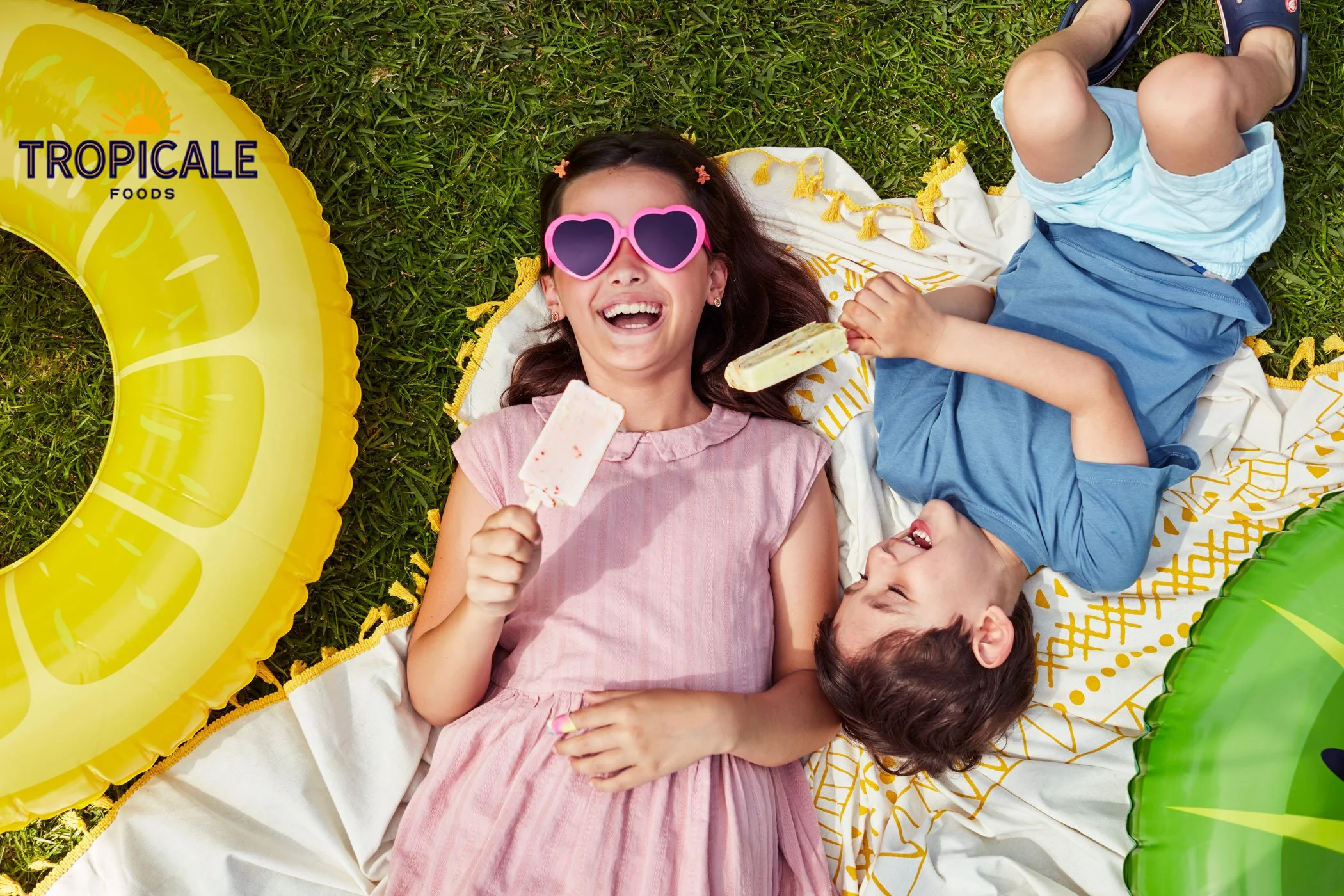 A girl wearing pink sunglasses and a pink dress is lying on a blanket with a boy wearing a blue shirt and blue shorts on a grassy lawn, surrounded by colorful inflatable fruit-shaped pool floats. The girl is holding a popsicle and smiling.