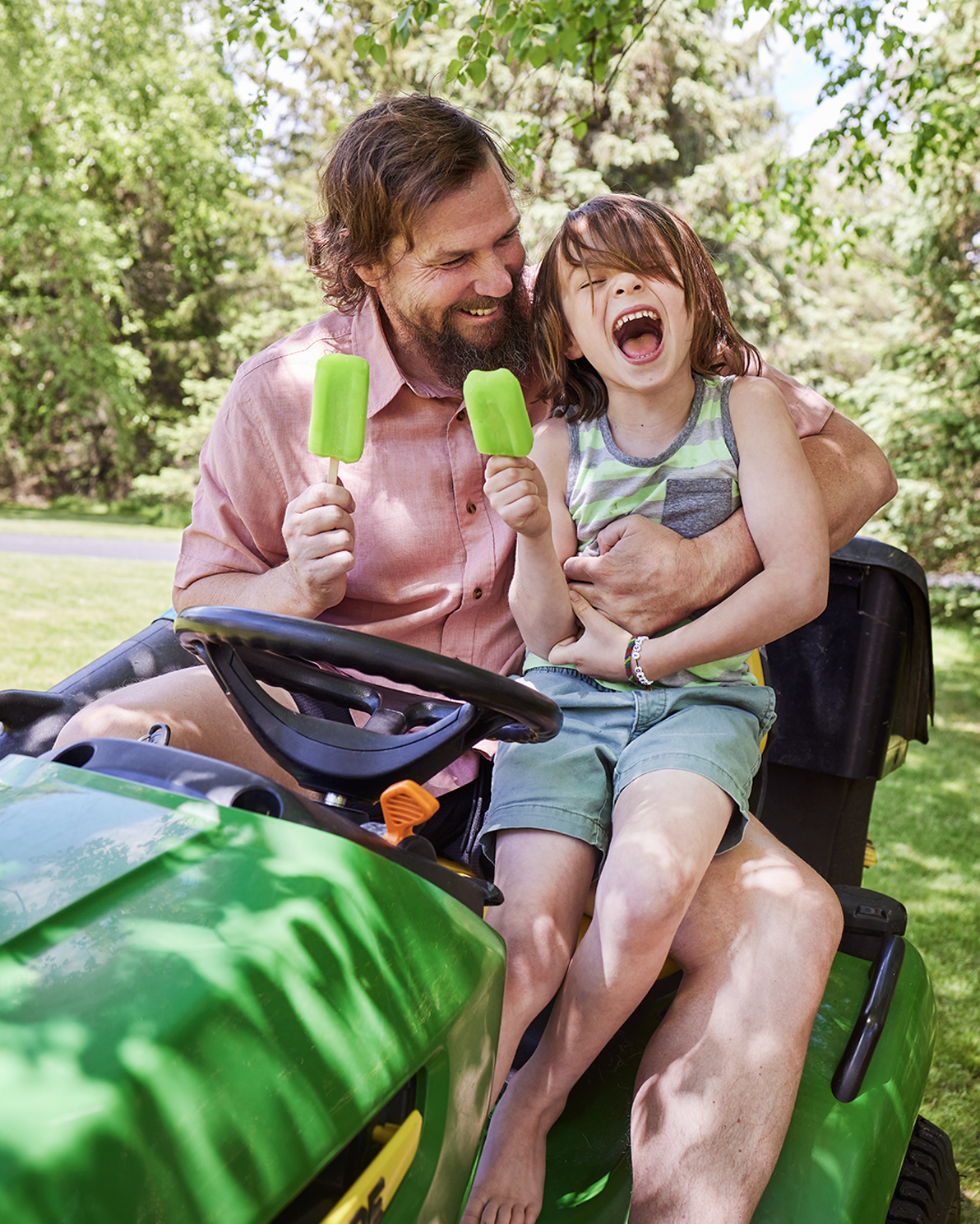 A man and a young girl sitting on a green riding lawnmower outdoors, enjoying ice pops and laughing.