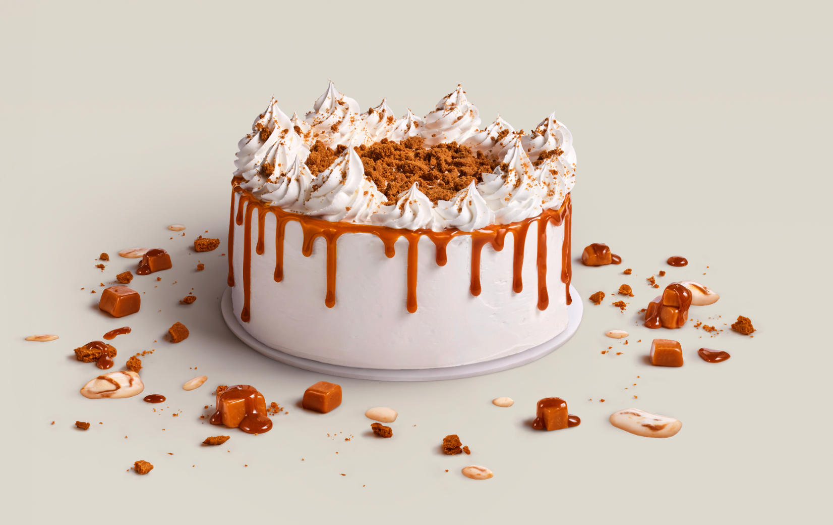 A round cake with white frosting, caramel drip, whipped cream swirls on top, crumbled cookies, surrounded by scattered caramel candies, cookie pieces, and caramel sauce on a plain light background.