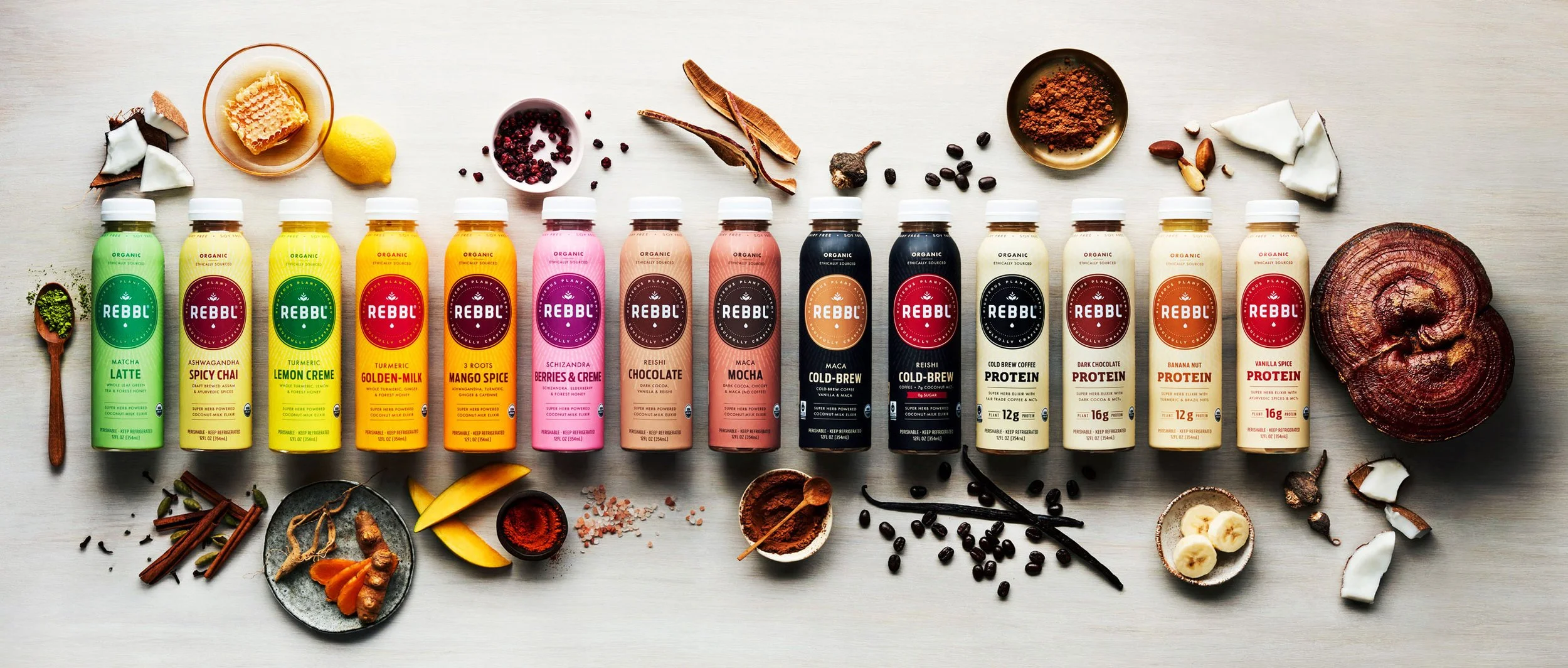 Array of 12 Rebb organic plant-based protein drinks in various flavors arranged in a line with ingredients like honey, lemon, spices, berries, chocolate, banana, and coconut scattered around them on a light-colored surface.