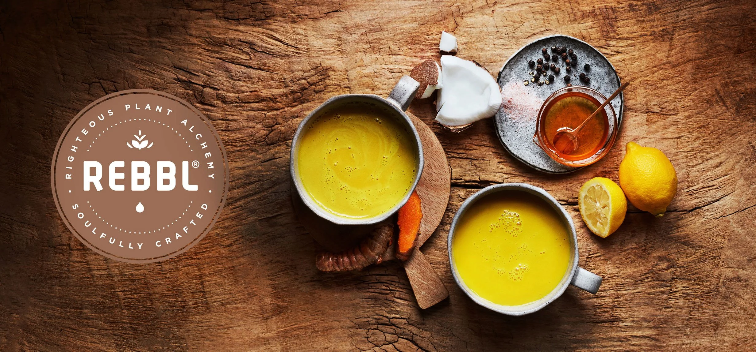 A wooden table with two mugs of yellow turmeric tea, a whole lemon and a lemon half, a small jar of honey with a spoon, a bowl of salt and pepper, and a piece of fresh coconut.