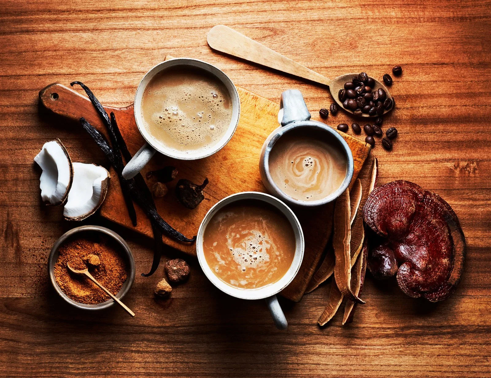 Three cups of coffee with foam on a wooden table, surrounded by vanilla beans, coconut, a small bowl of ground spices, coffee beans, and dried mushrooms.