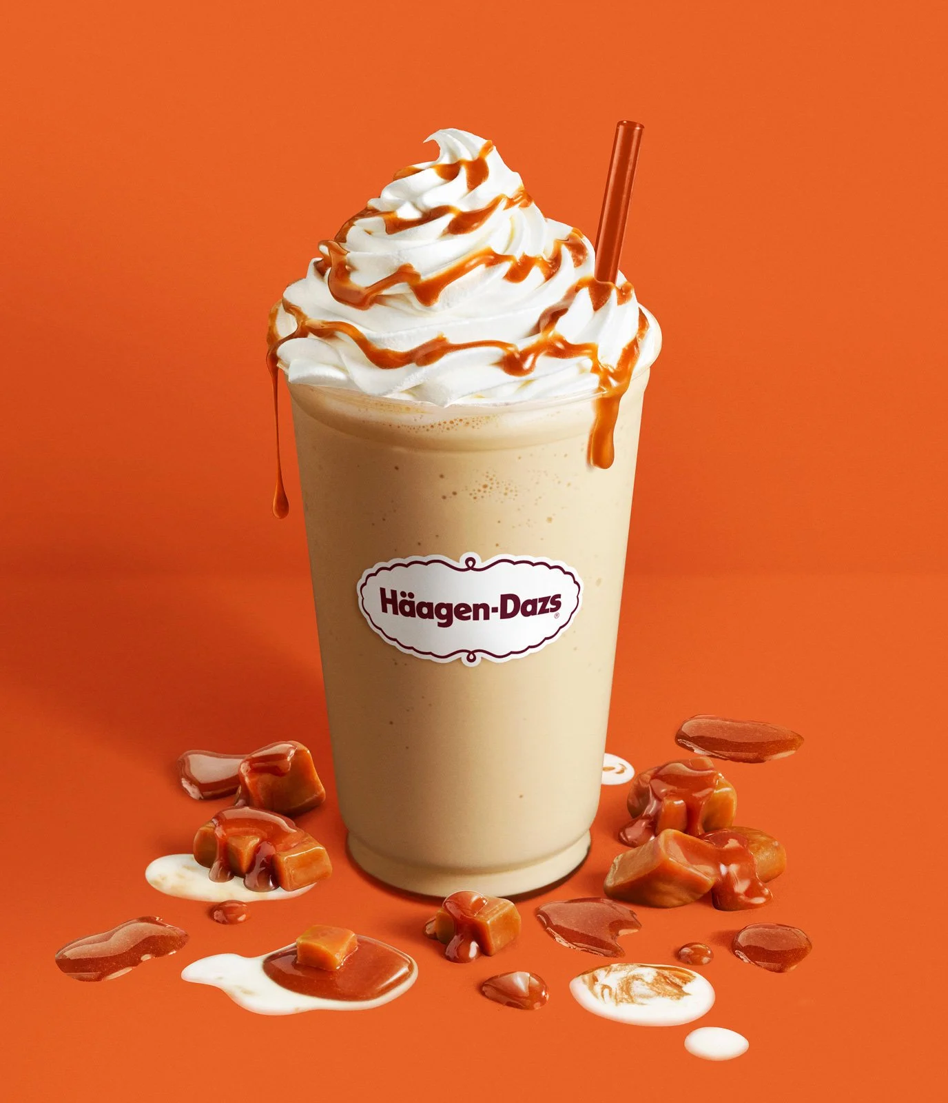 A caramel-flavored milkshake in a Häagen-Dazs branded cup, topped with whipped cream and caramel drizzle, with caramel candies around the base, on an orange background.
