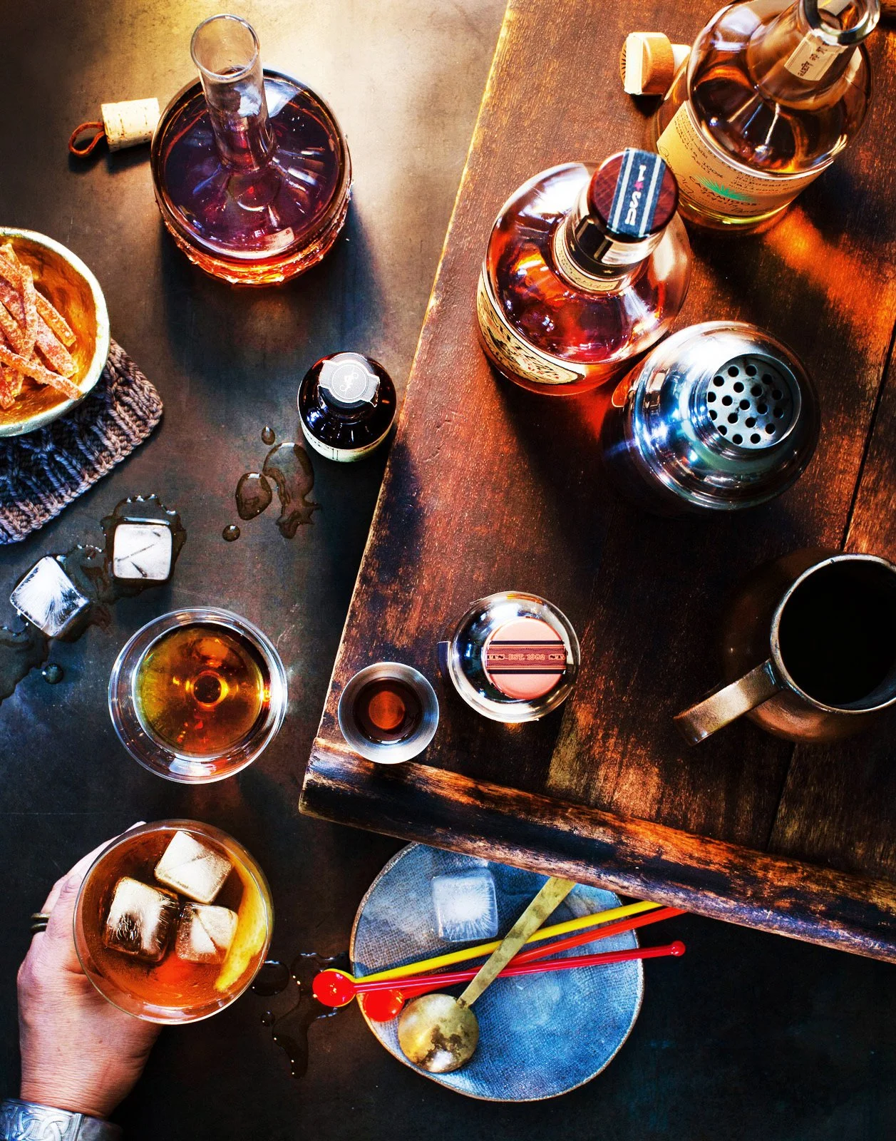 A top-down view of a bar scene with several bottles of alcohol, glasses filled with drinks and ice cubes, and bar tools on a dark wooden surface.