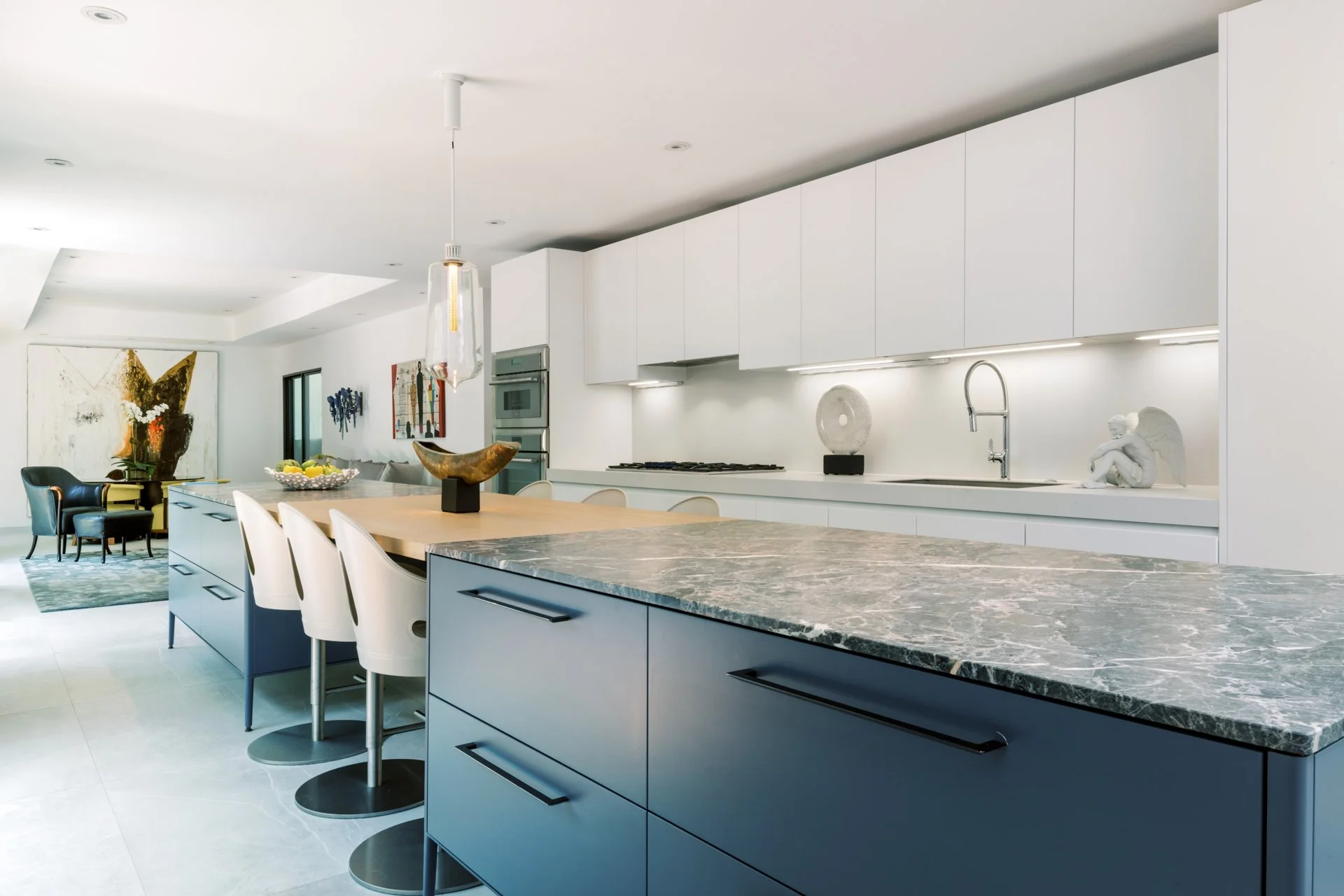 Modern kitchen interior with marble countertops, white cabinets, and a central island with seating. Features a pendant light, art pieces, and a sculpture.