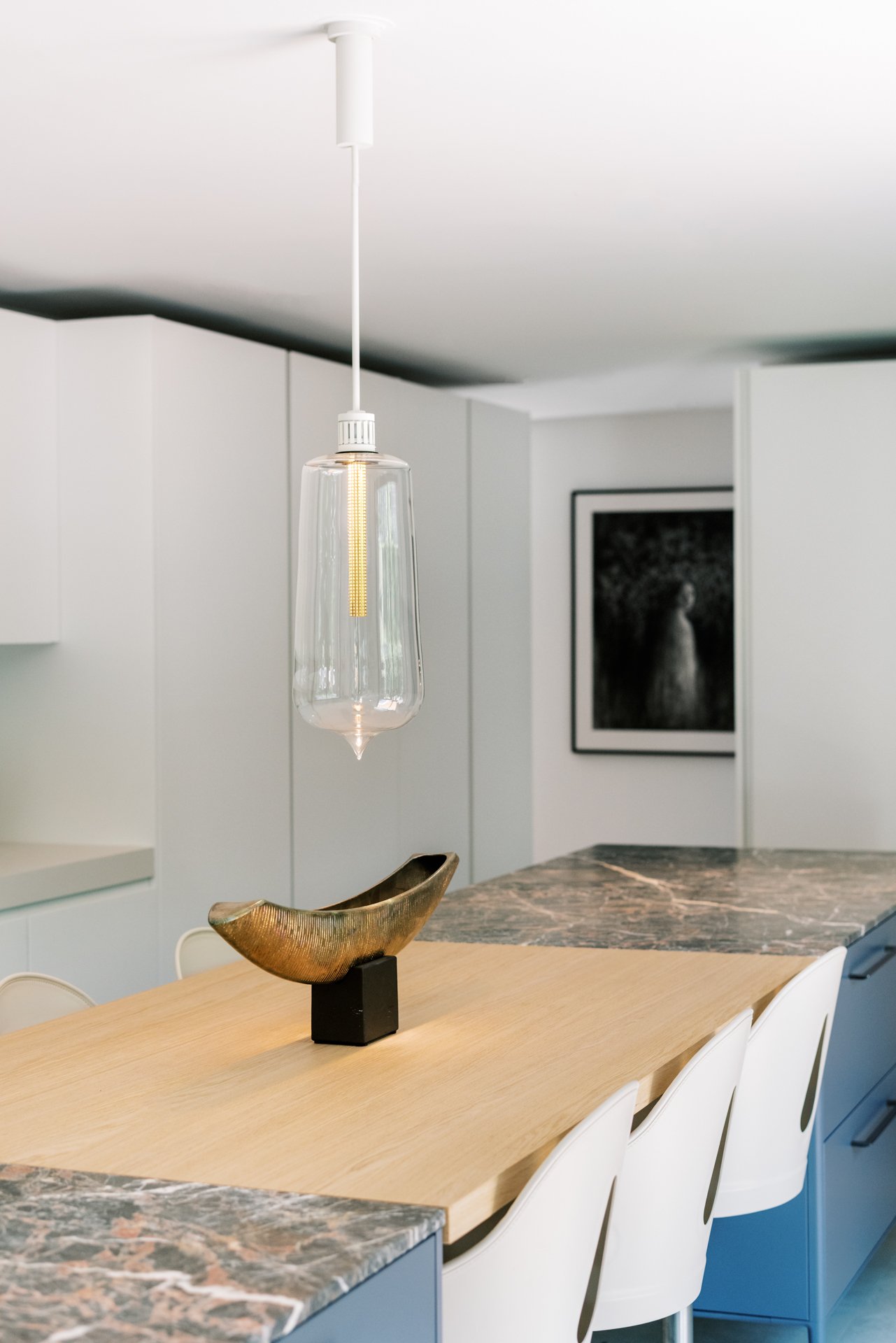 Modern kitchen with pendant light, wooden and marble island, white chairs, decorative sculpture, minimalist artwork on wall.