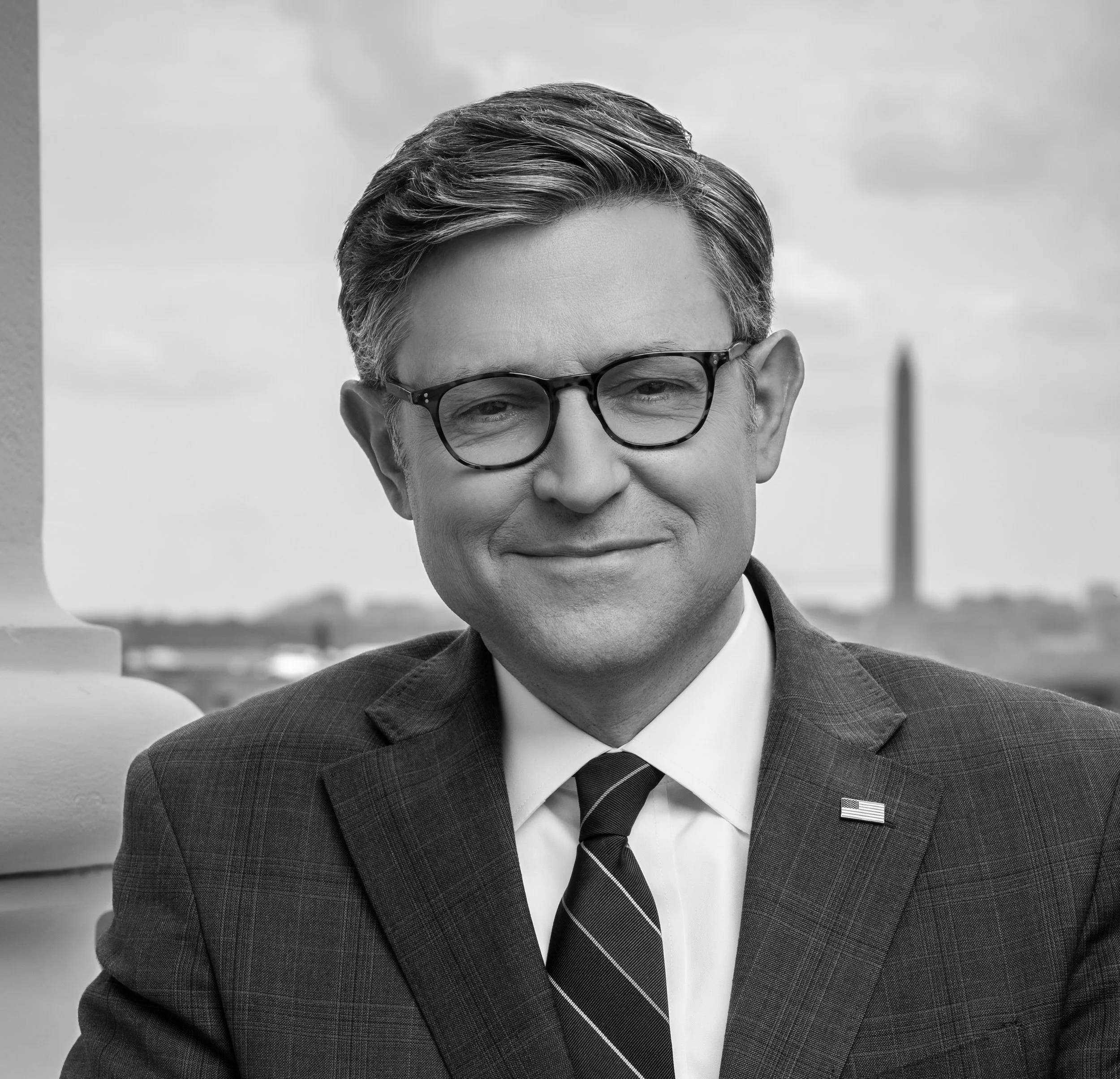 Black and white portrait of a man wearing glasses in a suit, outdoors with the Washington Monument in the background.