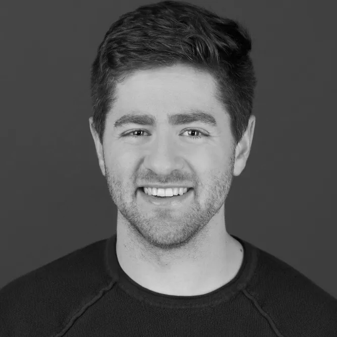 Black and white portrait of a smiling young man with short hair, light facial hair, wearing a dark shirt against a plain background.