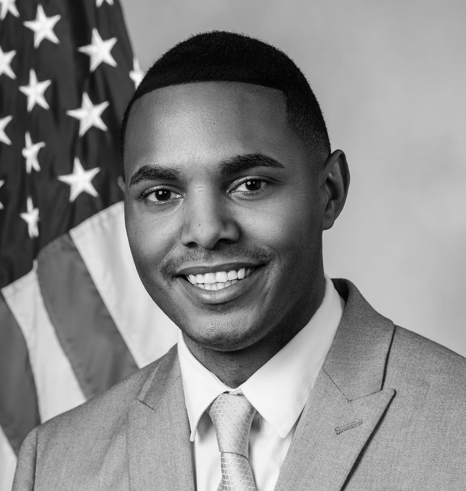 A professional portrait of a smiling African American man in a suit and tie, standing in front of a U.S. flag.
