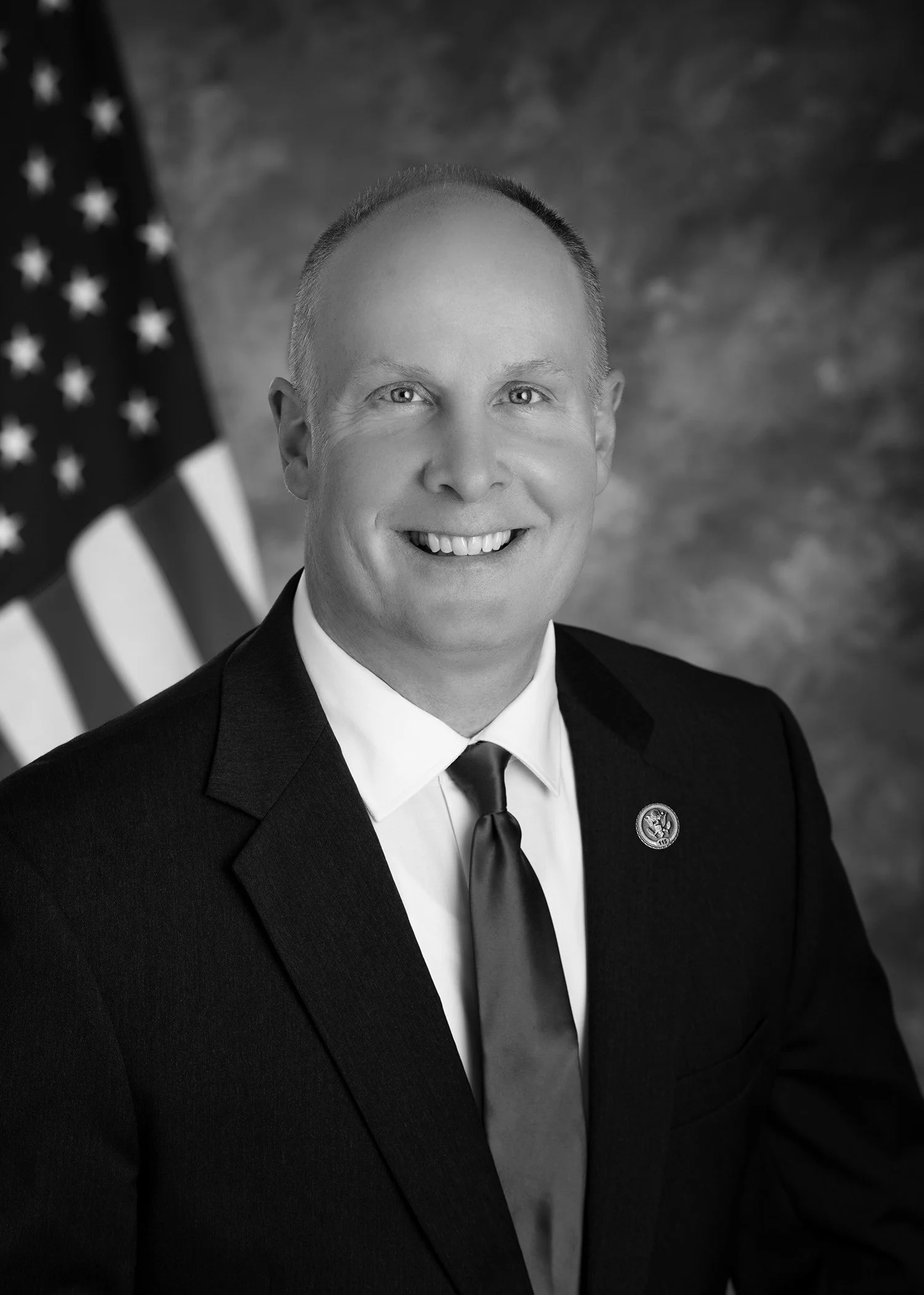 Black and white portrait of a smiling man in a suit, with an American flag in the background.