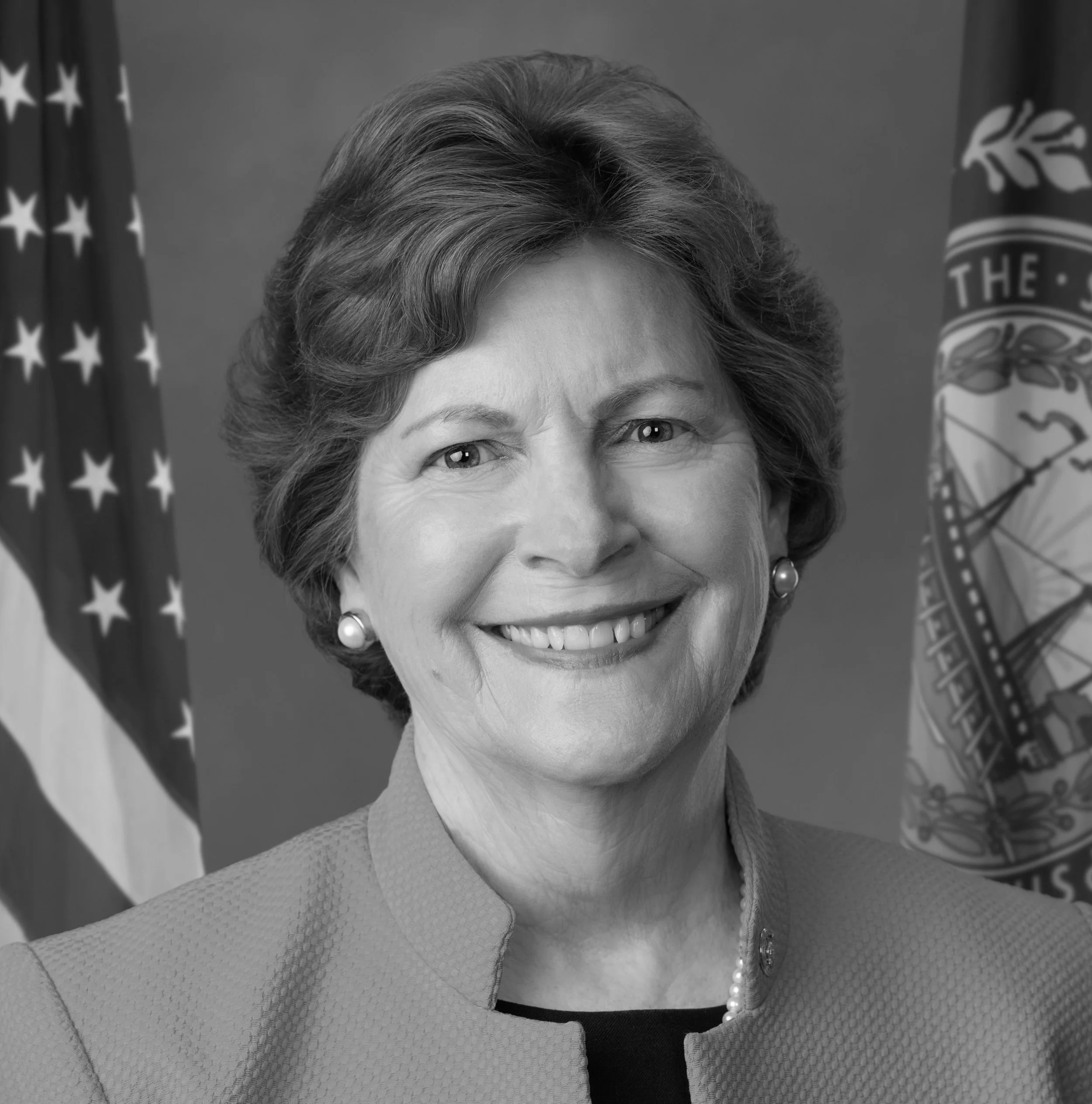 A black and white portrait of a smiling woman with short hair, wearing pearl earrings, a blazer, and a necklace, standing in front of two US flags.
