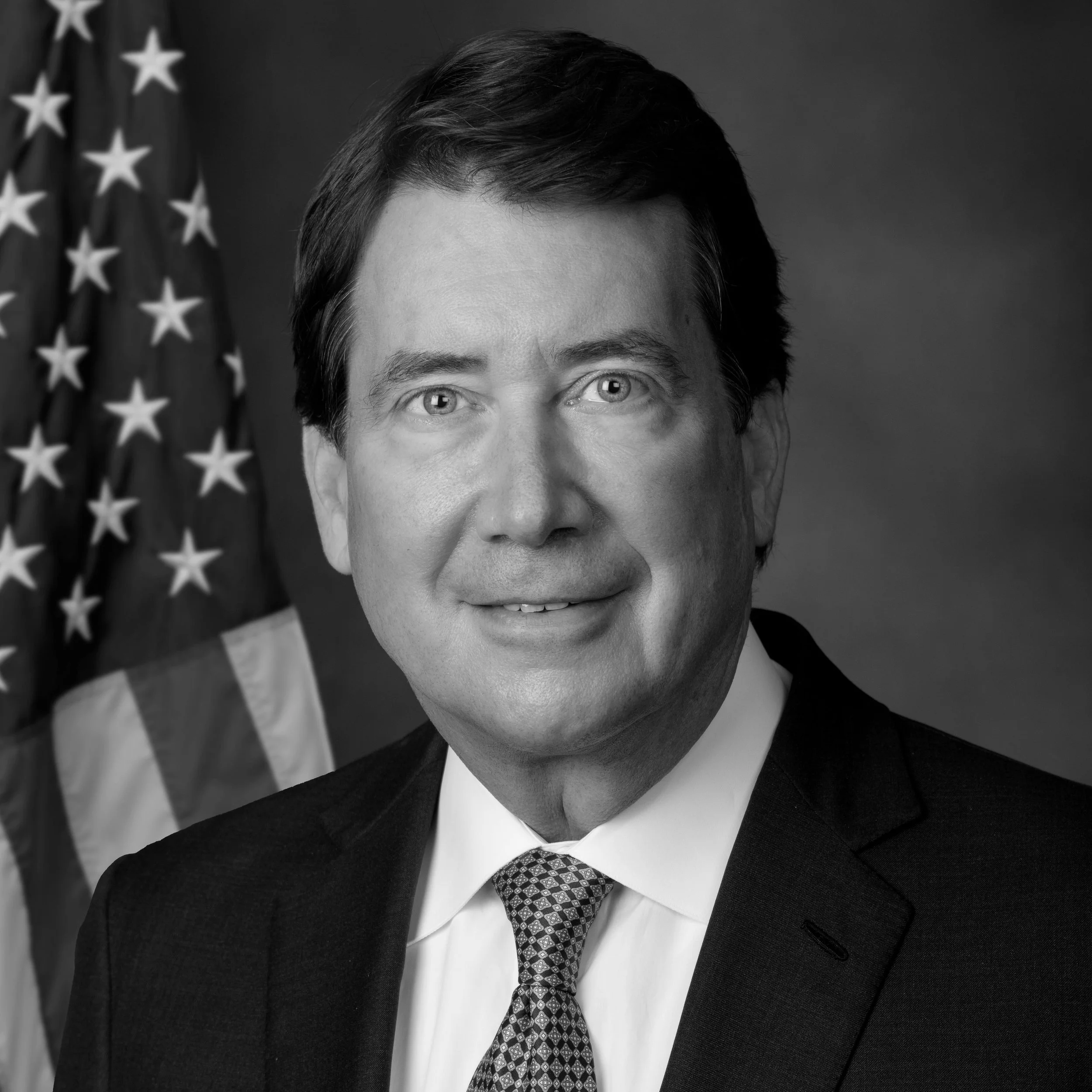 Black and white portrait of a man wearing a suit, tie, and white shirt, standing in front of an American flag.