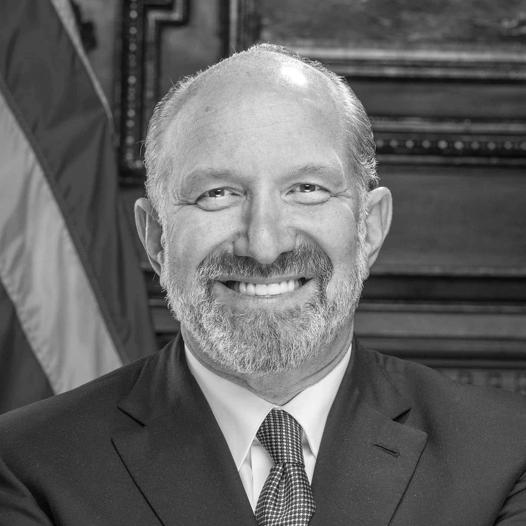 A black and white portrait of a smiling man with a beard, wearing a suit and tie, with an American flag in the background.