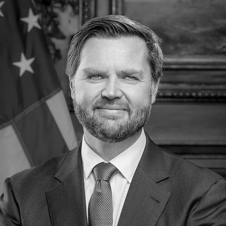 Black and white portrait of a man in a suit and tie, standing in front of an American flag, with a wooden wall in the background.