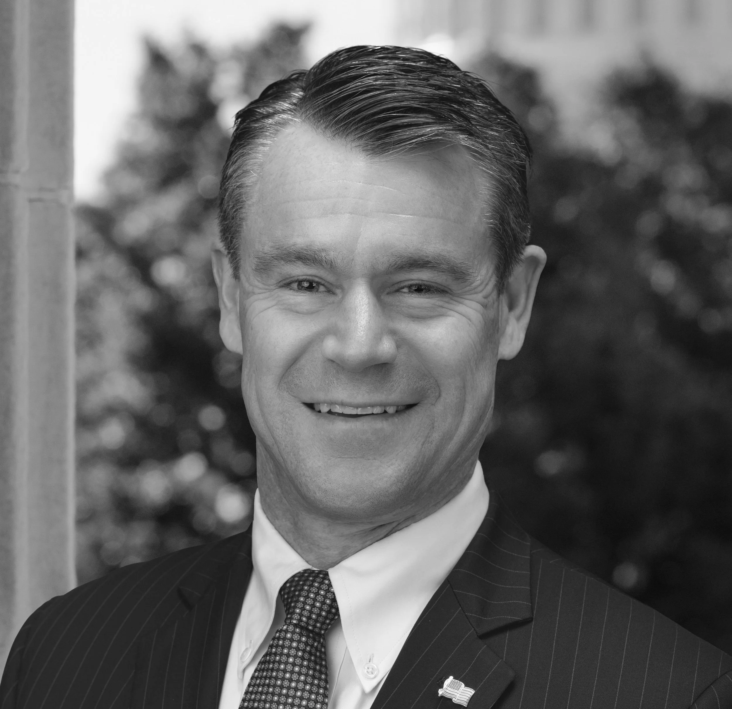 Black and white headshot of a smiling man in a suit, tie, and American flag lapel pin, standing outdoors with blurred trees in the background.