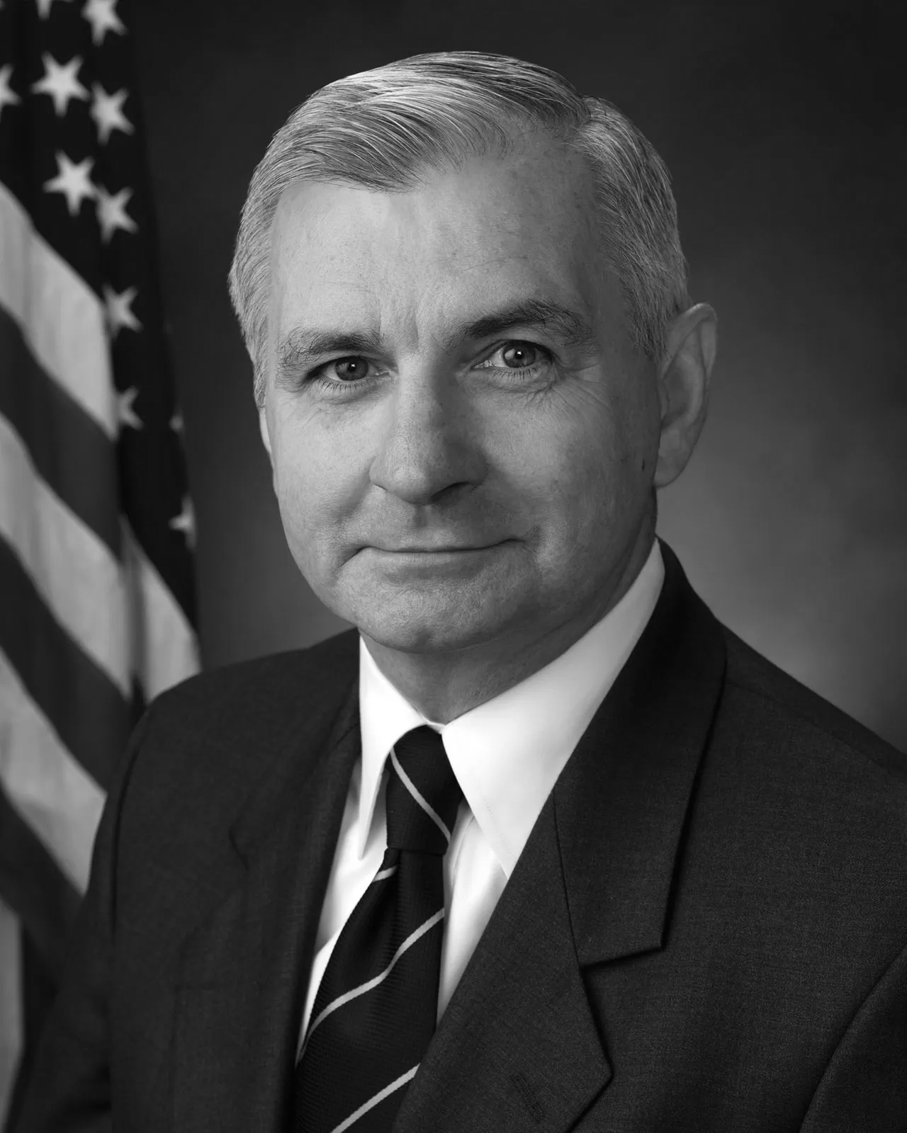 Black and white portrait of a man in a suit, with an American flag in the background.