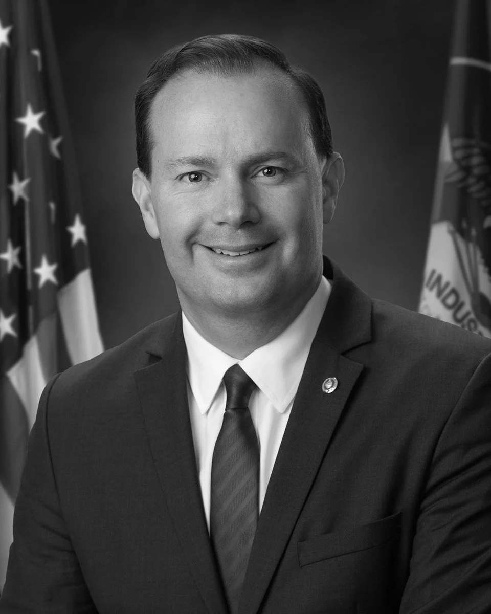 Black and white portrait of a smiling man in a suit, with flags in the background.