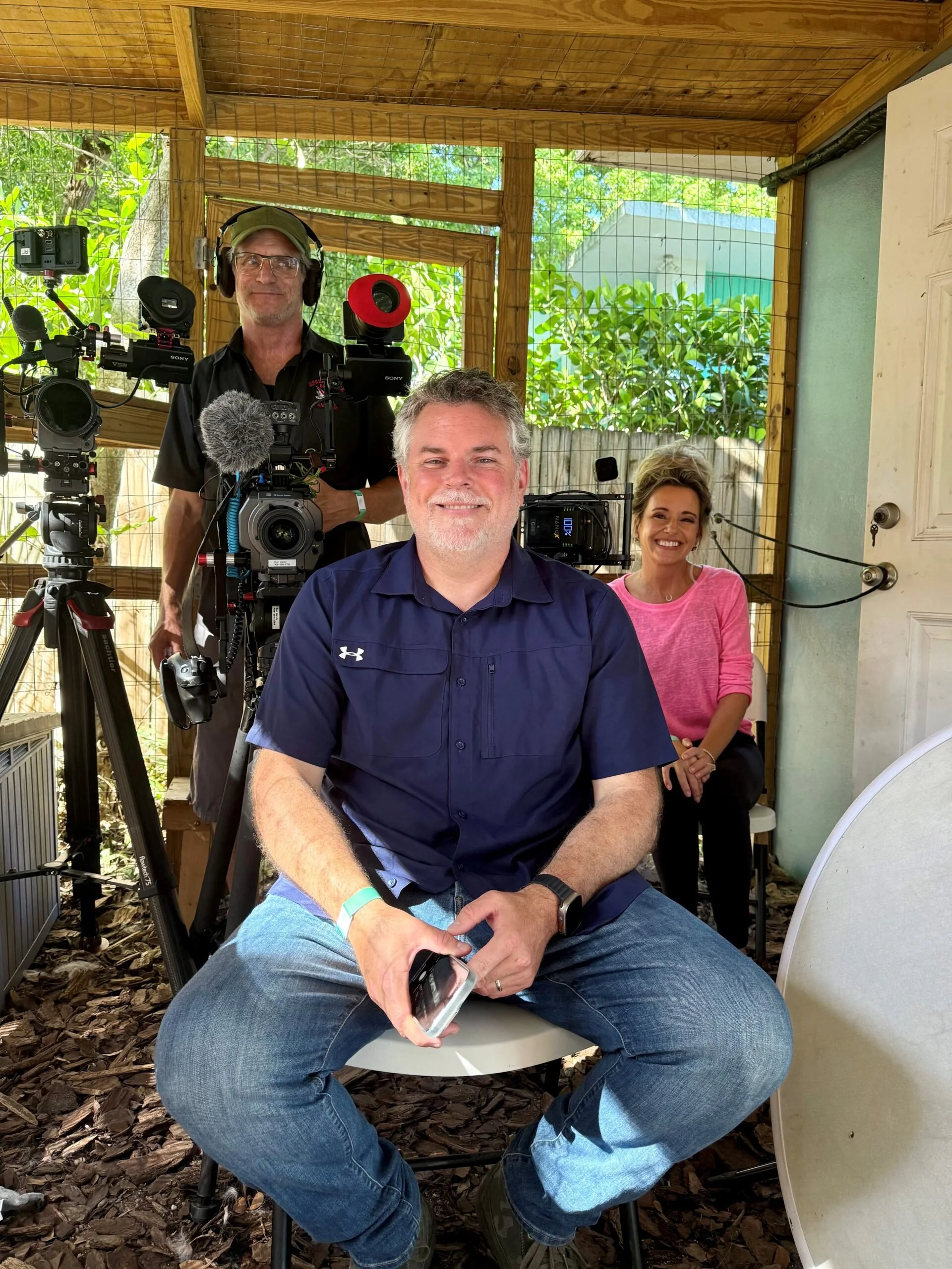 People in a filming setup with cameras and equipment, two men and a woman, inside a wooden structure with outdoor greenery visible.
