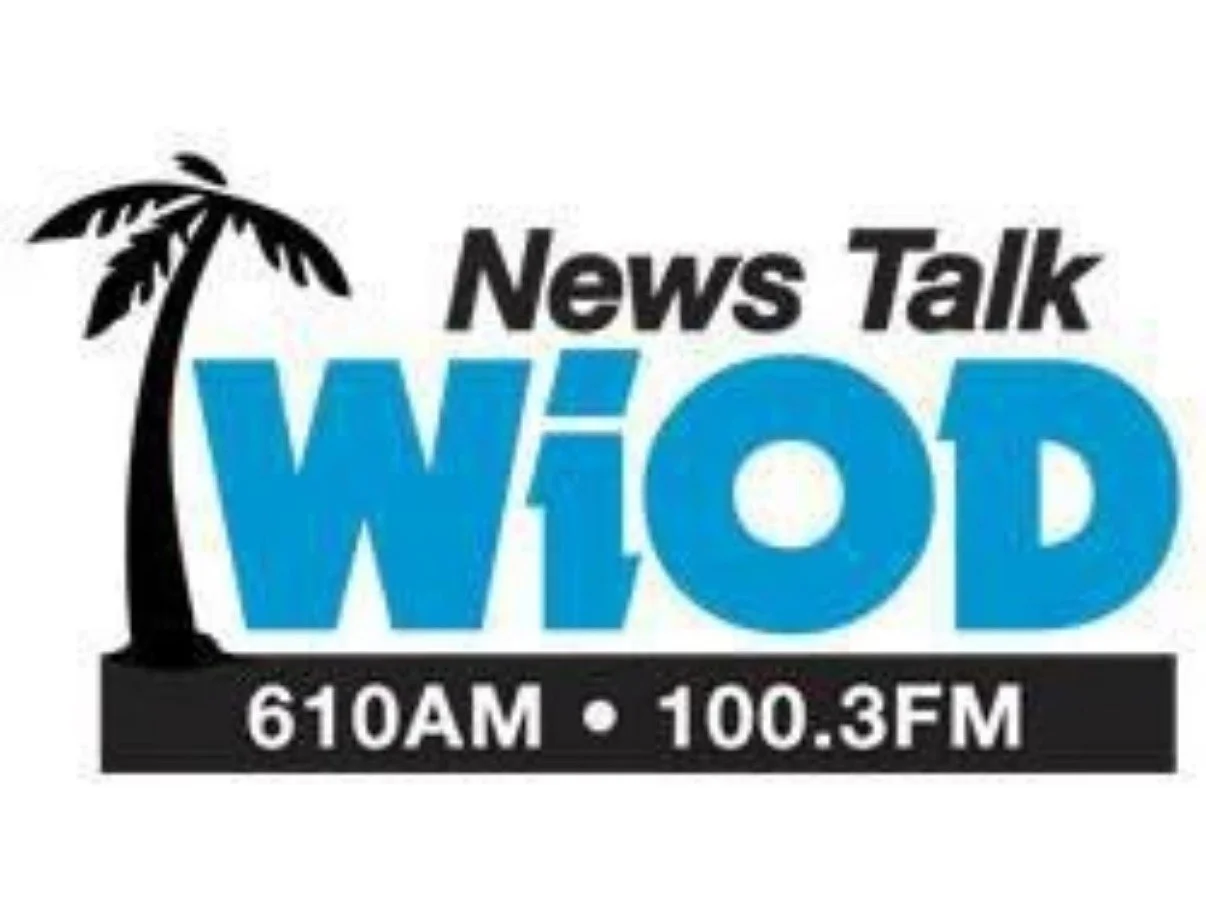 A promotional poster for a radio show feature with Jackie, showing her smiling at the camera with two dogs, a microphone, and a shelf with books and framed photos in the background. The poster includes the station logo WiOD, station frequencies, and a note about the show.