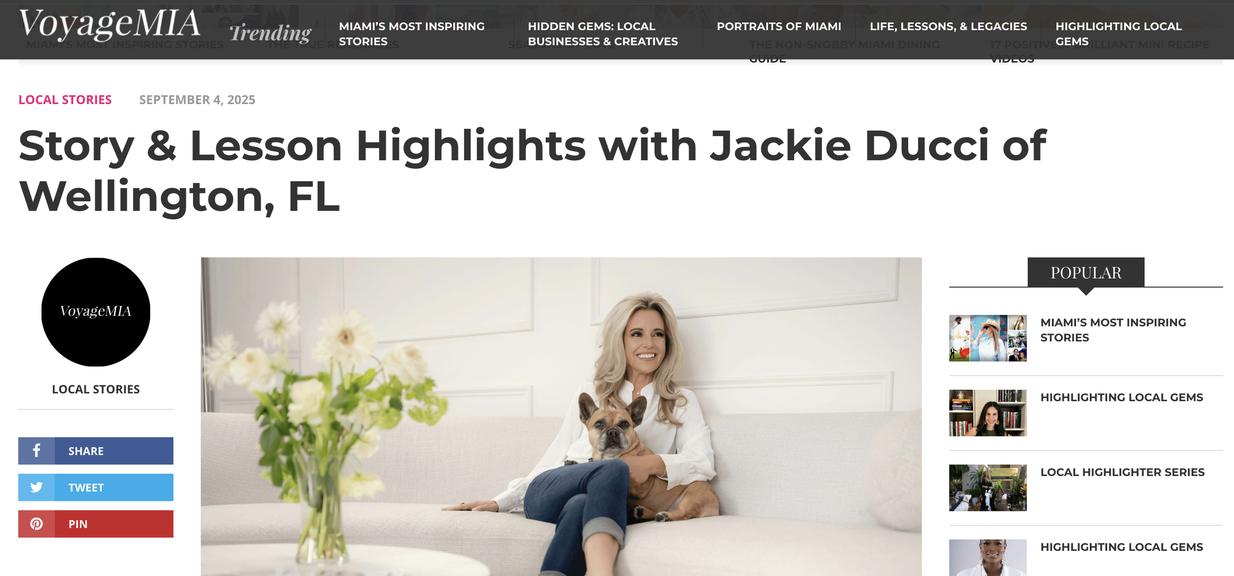 A woman with blonde hair, smiling, sitting on a white sofa with a brown and black French bulldog on her lap, in a bright room with white walls. A vase with white flowers is on a table in front of her.