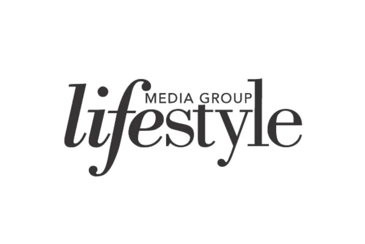 A woman with blonde hair smiling and hugging two bulldogs, sitting on a couch in a well-lit room with light-colored curtains in the background.