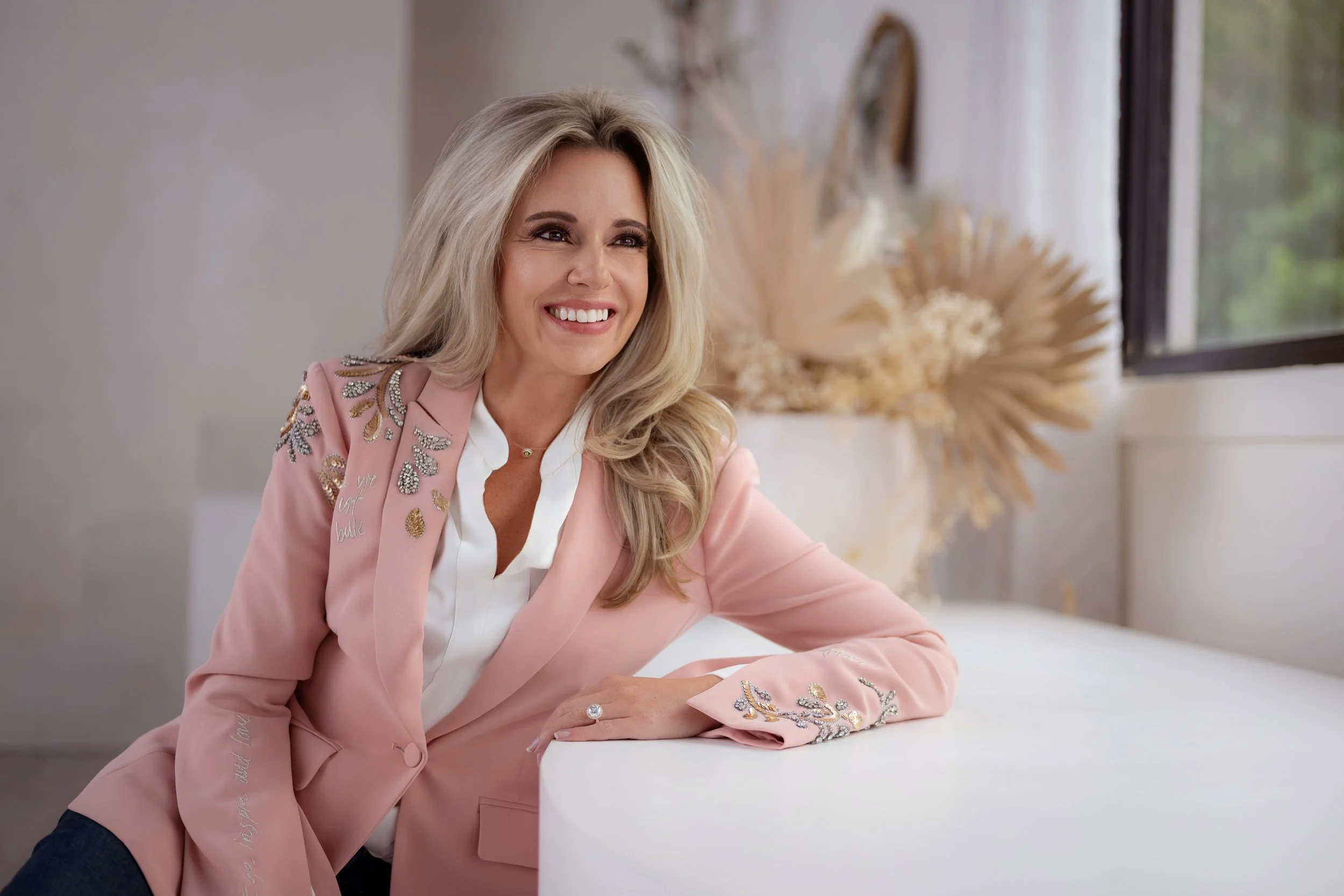 A woman with long blonde hair smiling while sitting at a white table in a bright room with large windows and dried plant decor in the background.