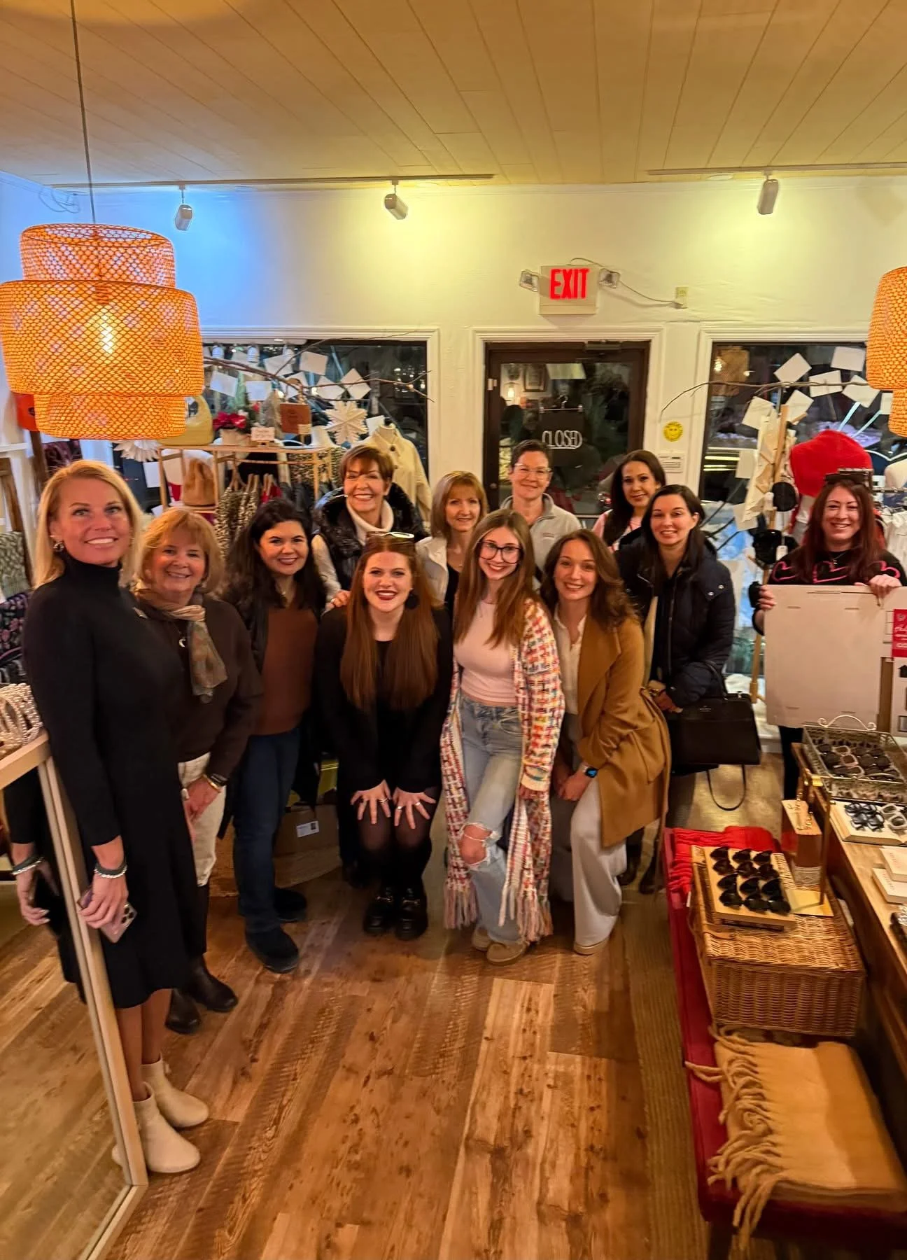 A group of eleven women standing together inside a store, smiling at the camera. The store has wooden floors, hanging orange lamps, and shelves with various items, including jewelry and accessories.