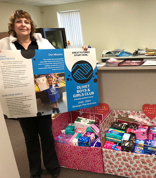 A woman standing inside an office next to two boxes filled with small wrapped gifts, holding a large informational poster about a community service project for the Olivet Boys & Girls Club.
