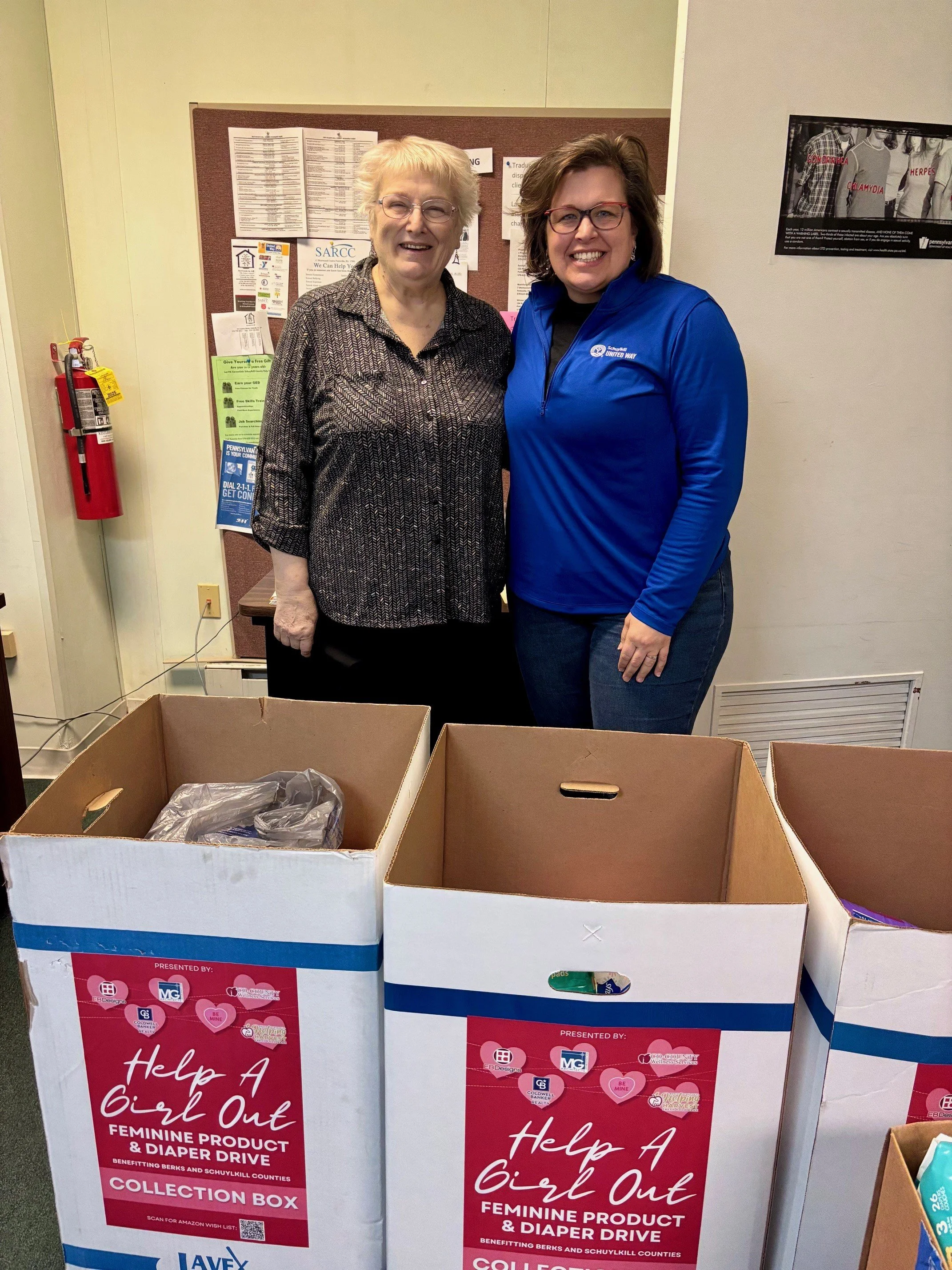 Two women standing behind donation collection boxes for a feminine product and diaper drive. The boxes have red and white posters with pink hearts, promoting the event to help girls out. One woman has short blonde hair and glasses, and the other has shoulder-length brown hair, glasses, and is wearing a blue jacket.