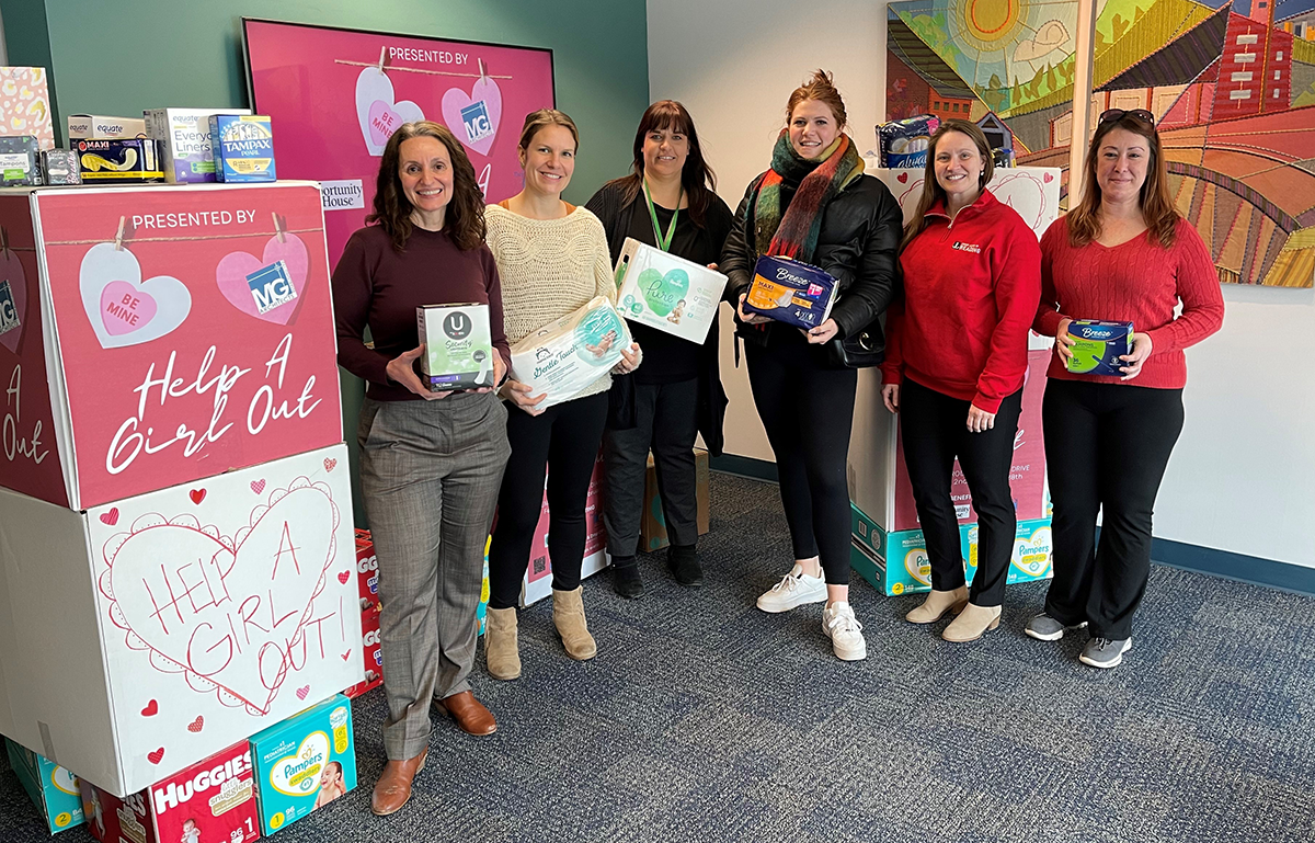 Six women standing in a room with a pink sign that reads "Help A Girl Out" and holding boxes of feminine hygiene products.