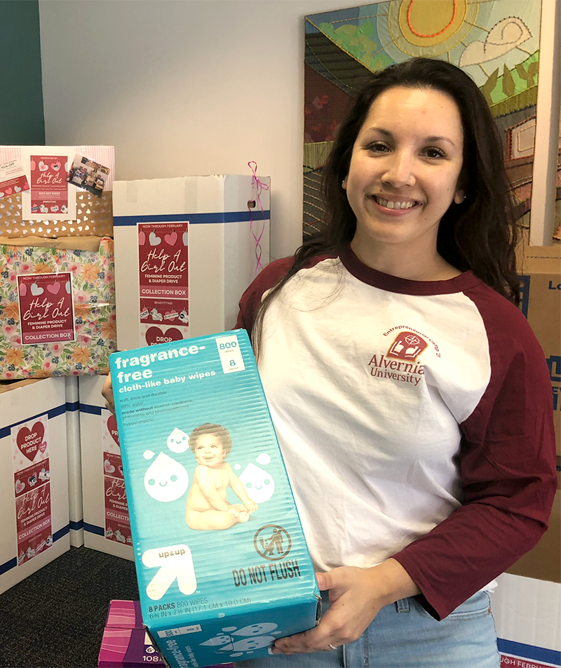 A young woman with long dark hair, wearing a white and maroon long sleeve shirt from Alvernia University, is smiling and holding a box of fragrance-free baby wipes. Behind her are boxes of donation items for a charity drive, with visible labeling for a collection box and gift bags. The background includes a colorful wall hanging.
