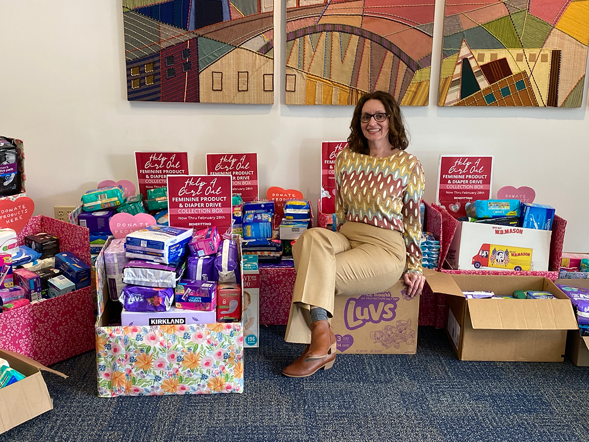 Woman sitting on a box of Luvs diapers in a room with donation boxes filled with feminine product and diaper collection items, signage indicates a drive benefiting women and children, colorful artwork on the wall in the background.