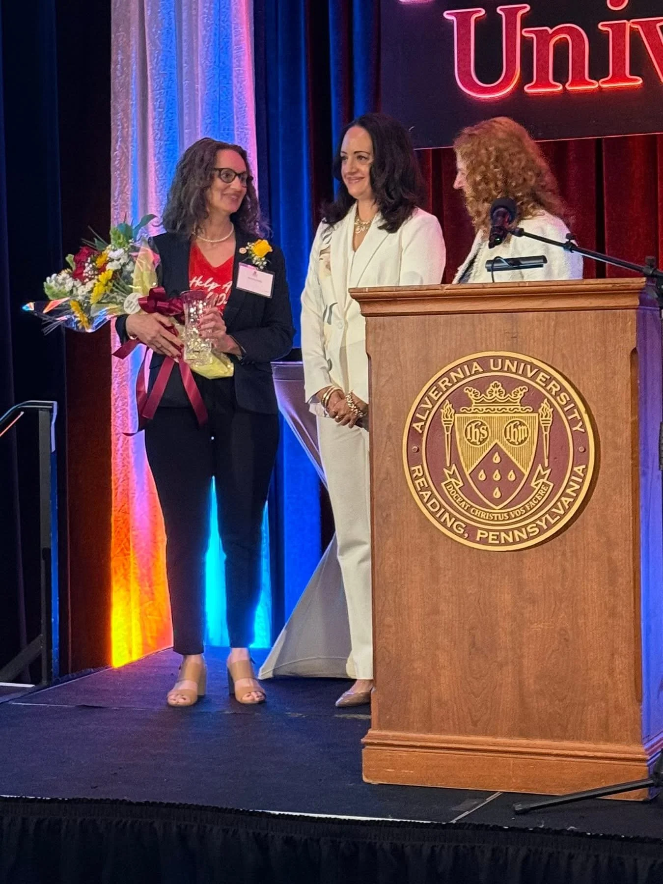 A woman holding a bouquet of flowers and a glass of champagne receiving an award on stage at a university event at Alvernia University in Reading, Pennsylvania. Two women stand nearby, one at a podium with the university seal. The background has colorful curtains and a partly visible red and black sign.