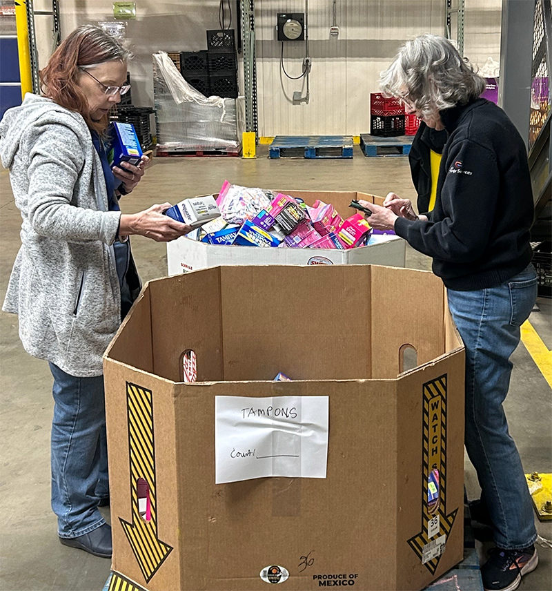 Two women are sorting tampons into a large cardboard donation bin in a warehouse. One woman is holding a box of tampons, and the other is using a smartphone. The bin is labeled "Tampons" and has a handwritten note and warning labels.