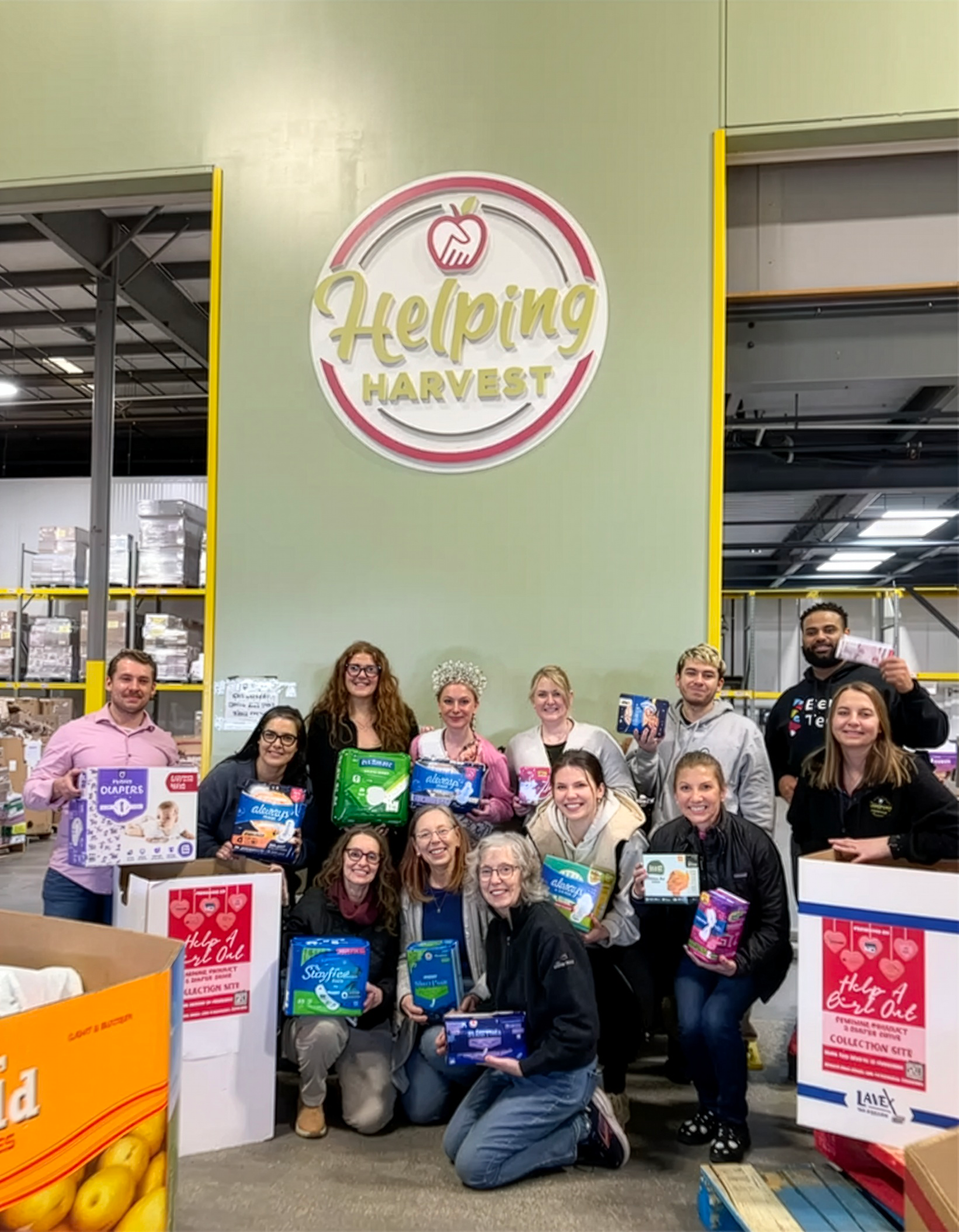 Group of people at a charity event inside a warehouse, holding feminine hygiene products, with a large "Helping Harvest" sign in the background.