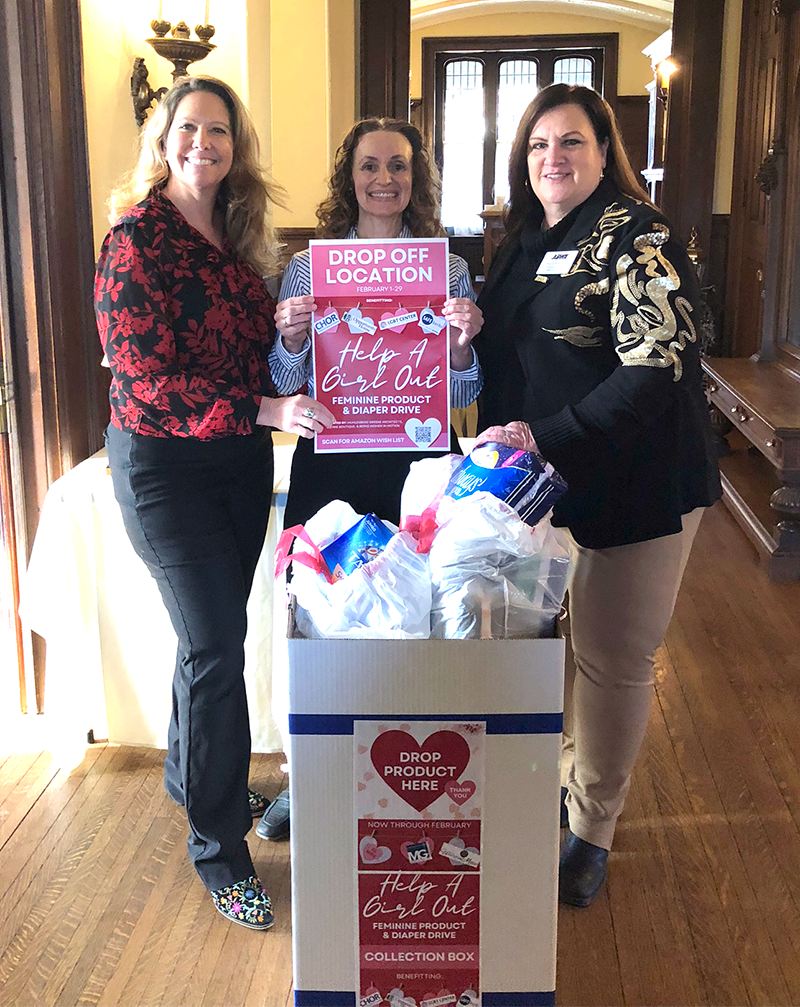 Three women standing around a donation box filled with feminine products and diapers at a collection drive event. One woman holds a sign that says Help A Girl Out for a feminine product and diaper drive.