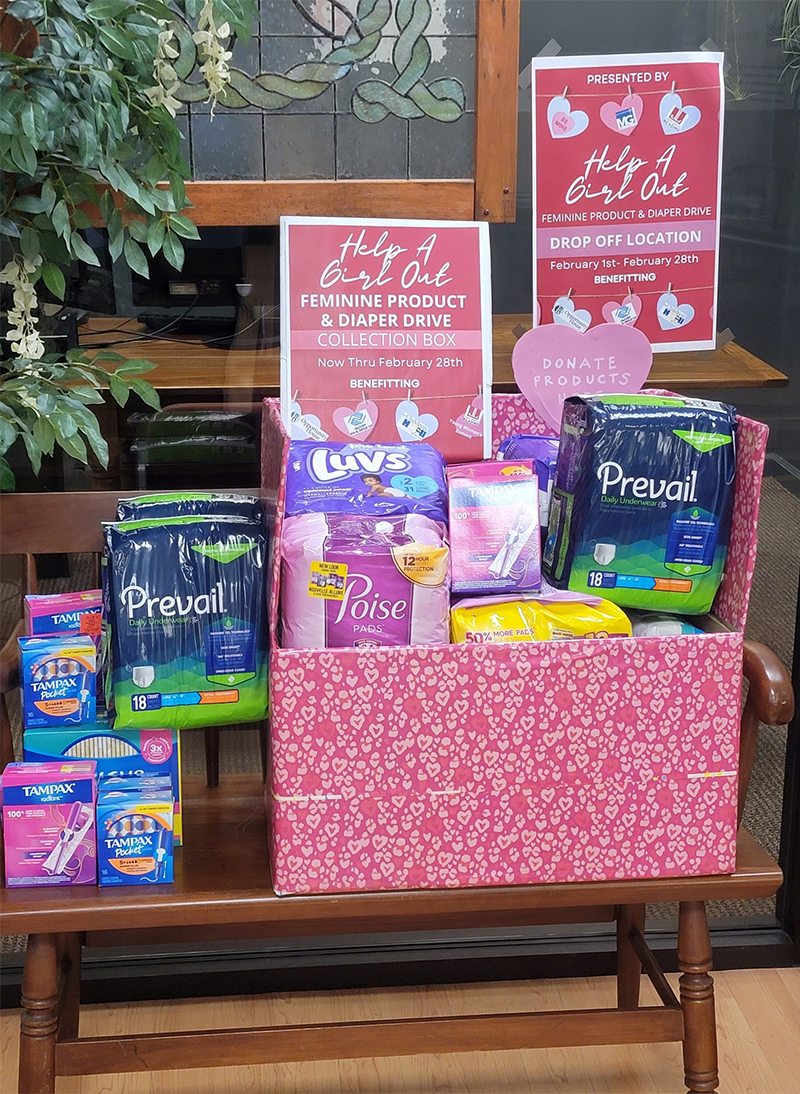 A pink box filled with feminine hygiene products, including packages of Prevail adult underwear, Poise pads, and Tampax tampons. There are signs indicating a 'Help A Girl Out' drive for feminine products and diaper donations.