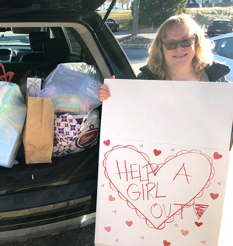 A woman with blonde hair and sunglasses standing by an open car trunk, holding a large white sign with a red heart and the words 'Help a Girl Out' written inside it, surrounded by bags and items inside the trunk.