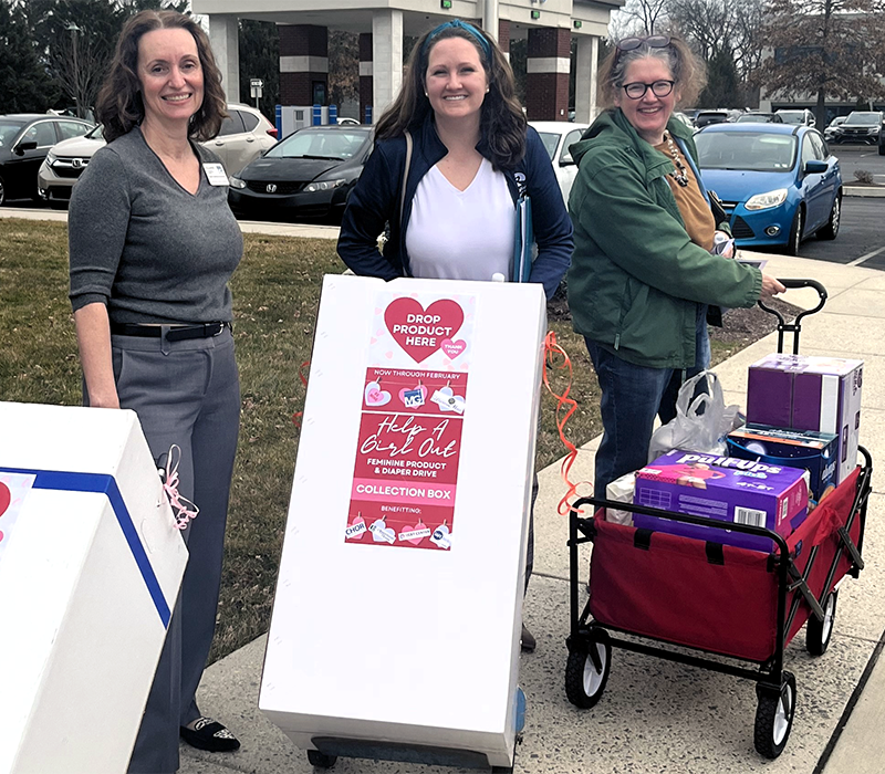 Three women standing outside near a donation collection box and a shopping cart filled with boxes. The woman in the middle is holding a sign promoting a feminine product and diaper drive to help girls out.