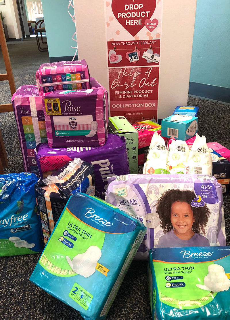 Display of various feminine hygiene products, including pads and adult diapers, in front of a pink poster urging donation of feminine products for a charity drive.