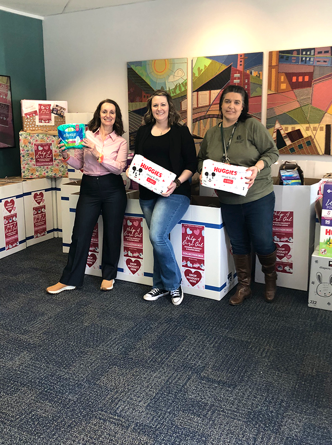 Three women standing inside a room, holding boxes of Huggies diapers, in front of donation boxes filled with baby supplies, with colorful abstract artwork on the wall behind them.