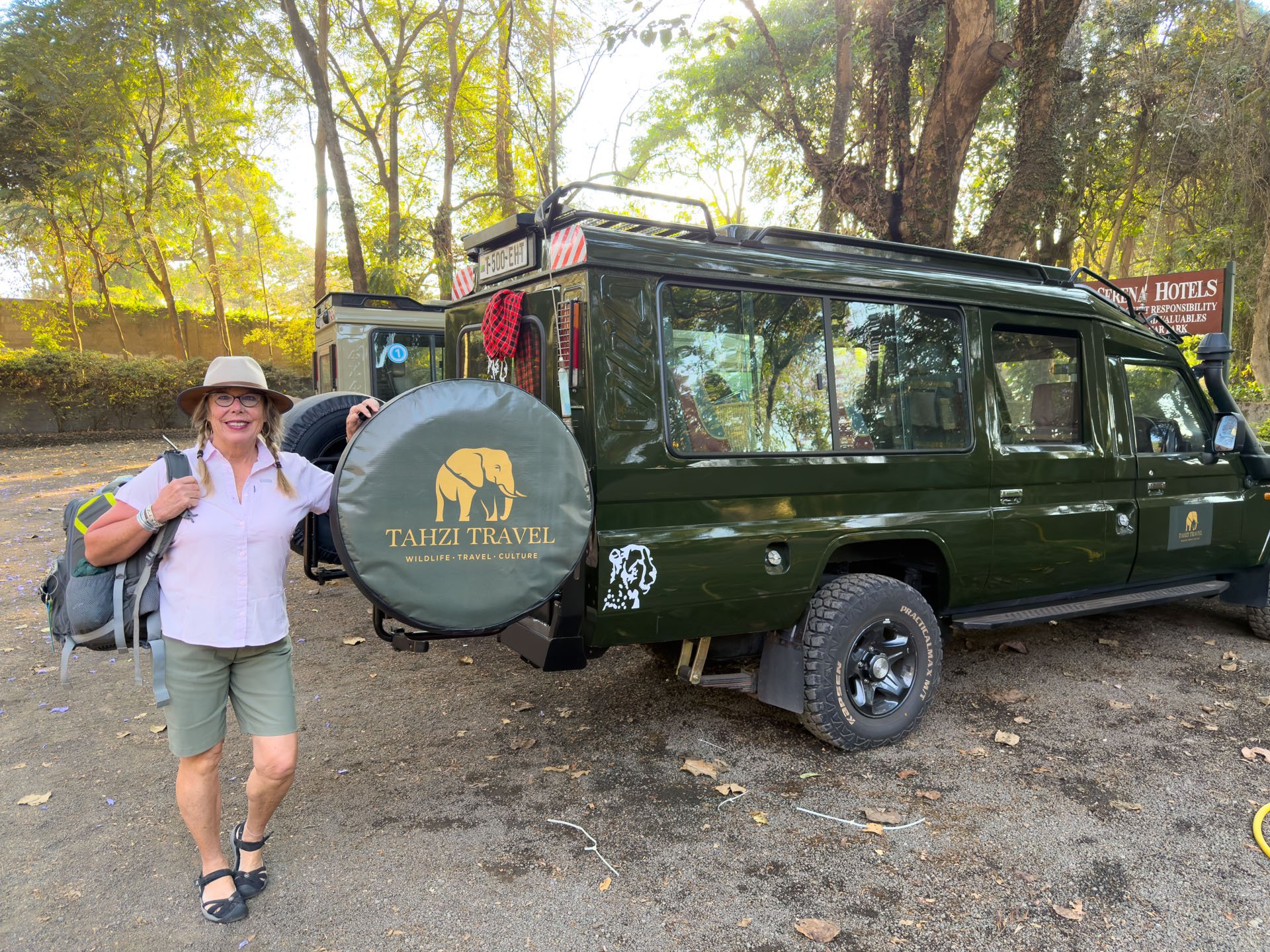 Co-founder of 'Tahzi Travel', Linda, Banaszak smiling, wearing a hat, glasses, pink shirt, and khaki shorts, with a backpack, in a wooded area with trees and signs in the background.