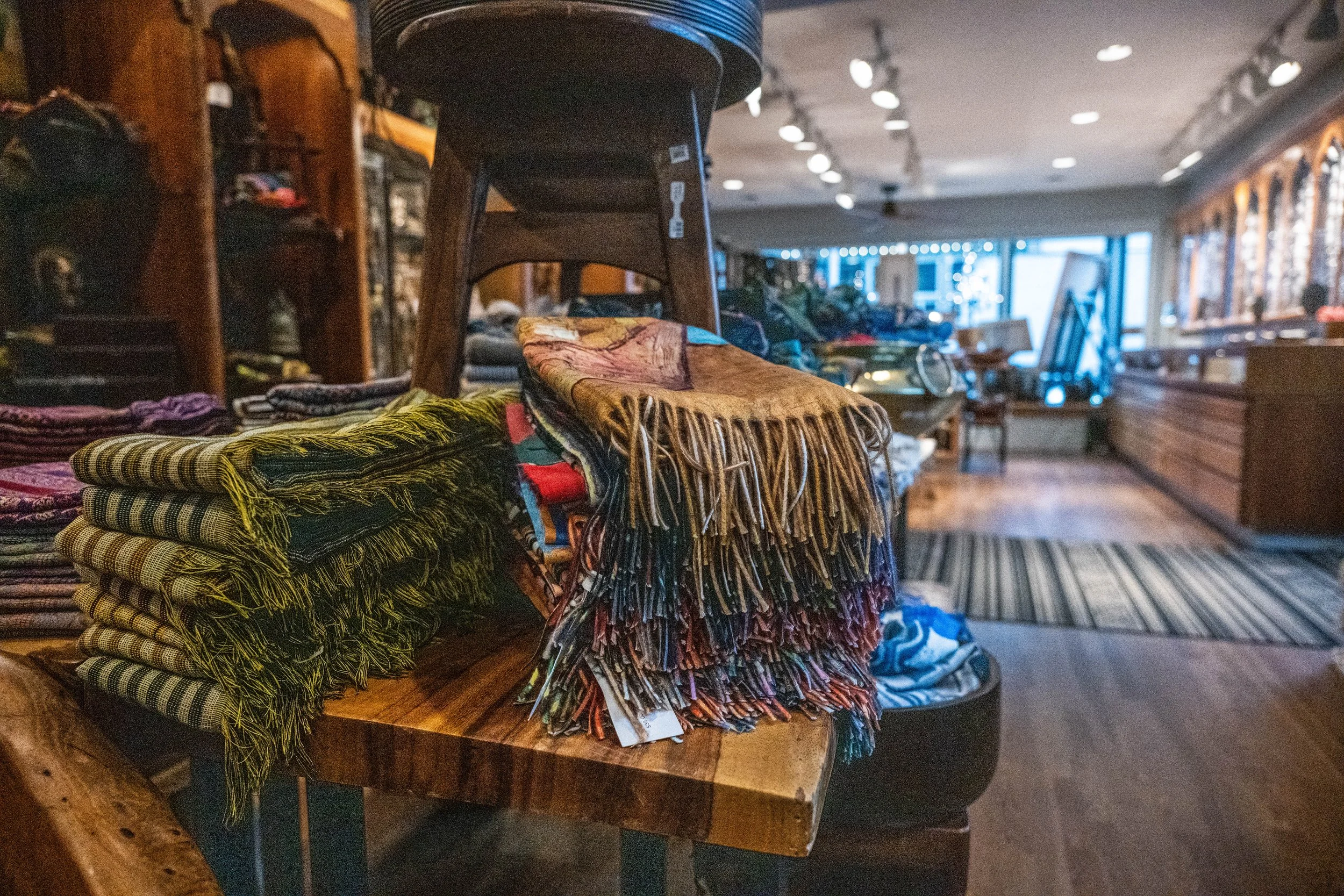 Caravan Gallery in La Conner, WA -  Stacks of folded patterned scarves and a woven scarf with fringes on a wooden table inside a vintage shop, with display shelves and a counter in the background.