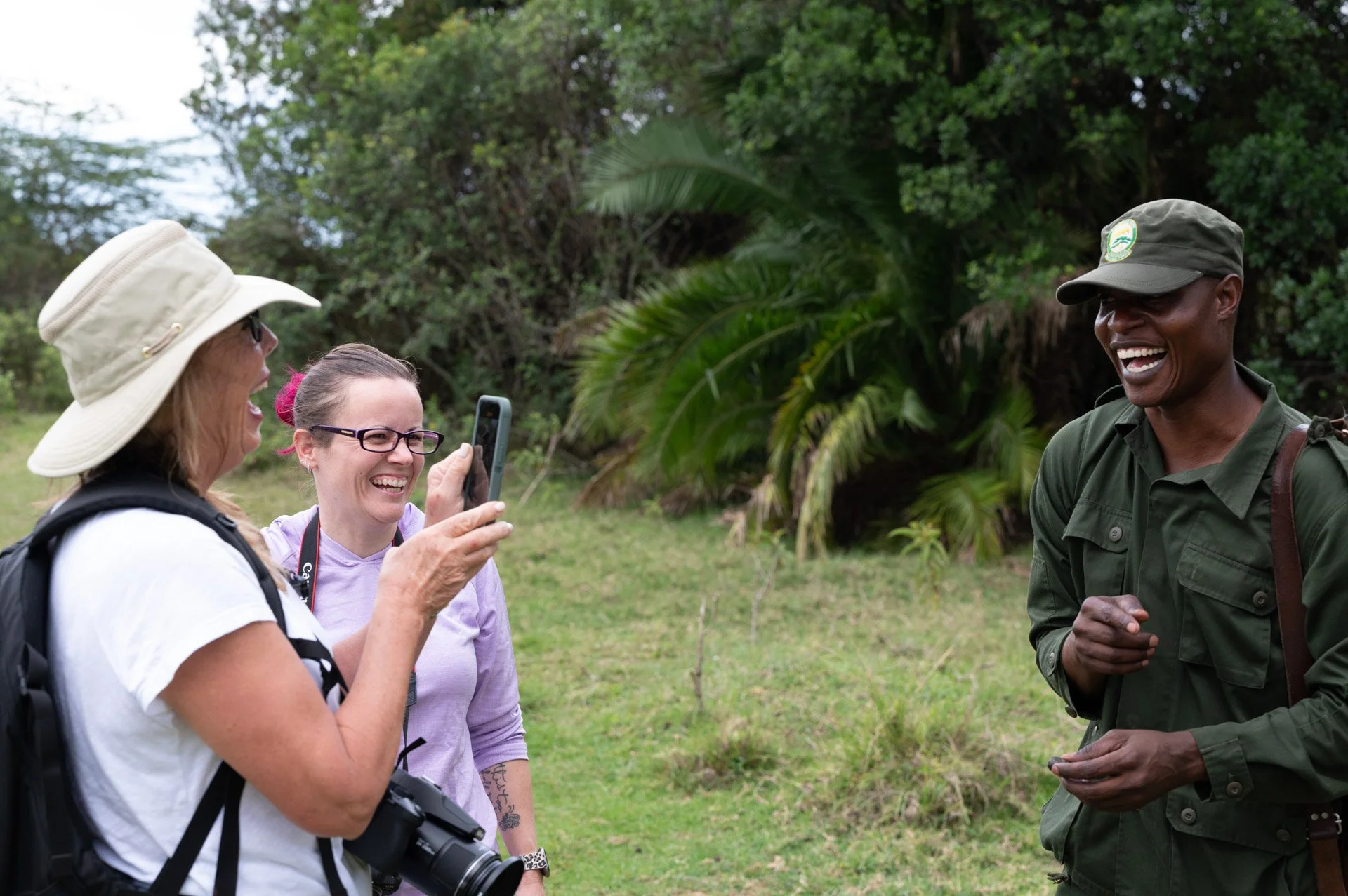 Caravan Gallery in La Conner, WA - Owner Linda Banaszak  chat and laugh with a park ranger in uniform during a sunny day in a grassy area with trees and bushes. Arusha National Park, Tanzania. 