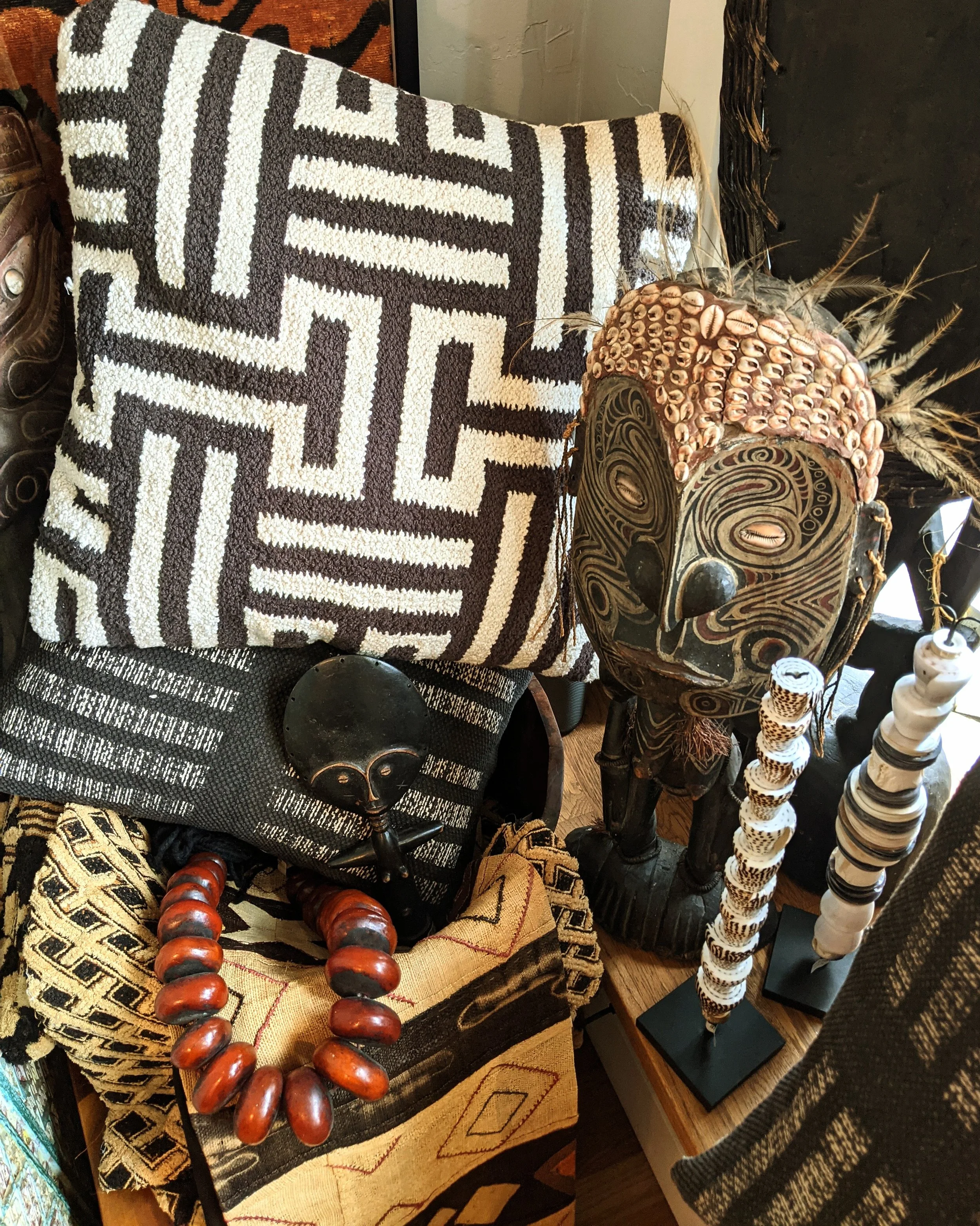 A display of African tribal artifacts including a large wooden mask with intricate swirling carvings, decorated with cowrie shells and feathers; a smaller black, carved wooden mask with exaggerated facial features; a necklace with large polished red beads; patterned textiles with geometric designs in black, white, and beige.