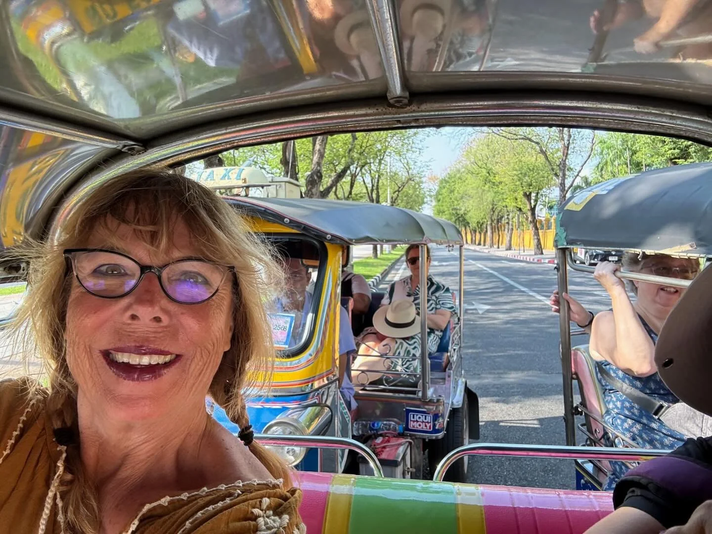 A woman with glasses smiling inside a tuk-tuk, with other passengers visible in the background, on a tree-lined street in daytime.