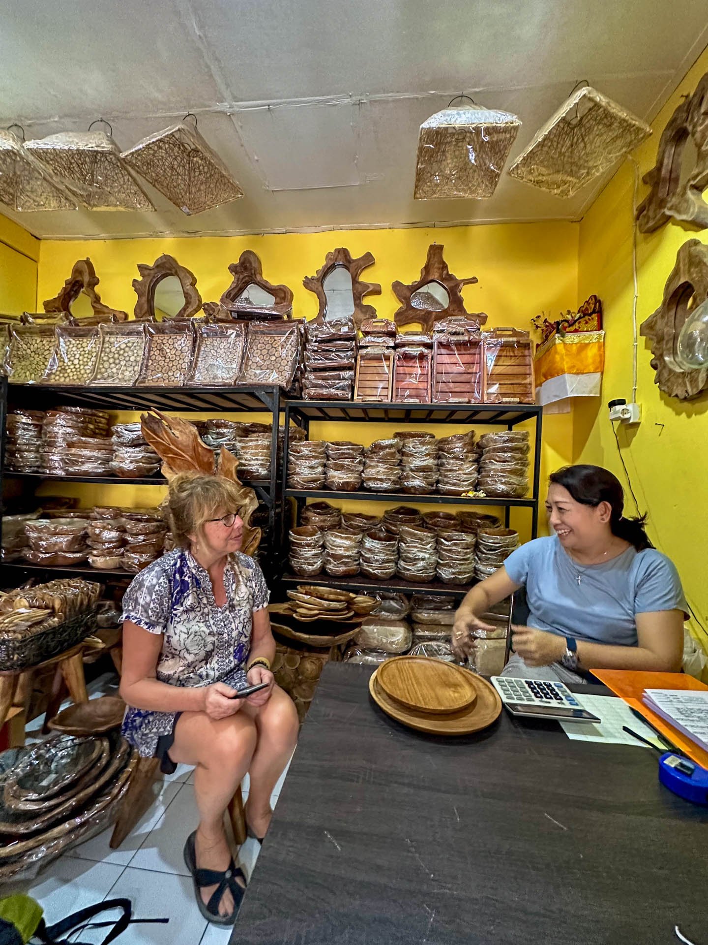Two women sitting in a shop surrounded by wooden crafts and shelves filled with wooden trays and decorative items, engaging in a conversation with smiles. Available at Caravan Gallery in La Conner, WA - 