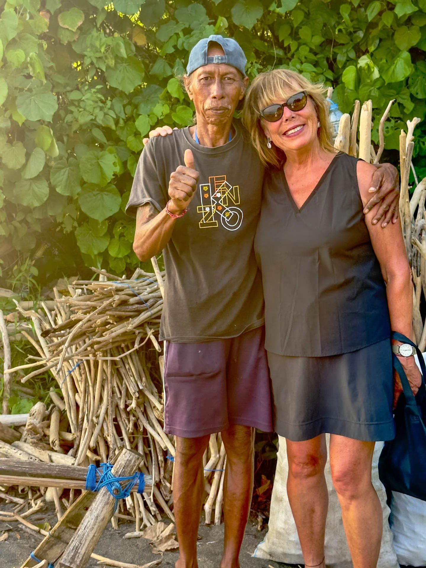 Two women standing next to each other outdoors, one giving a thumbs-up gesture, with greenery and a pile of sticks in the background.