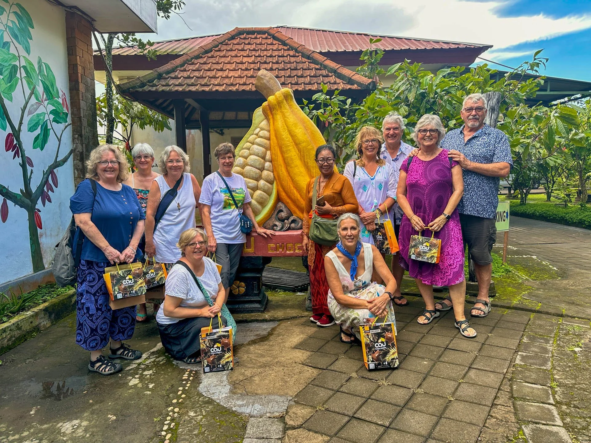 A group of eleven women and one man posing outdoors in front of a large giant cocoa pod sculpture at a cacao farm or botanical garden. Some women are holding gift bags with cacao or chocolate branding. The background includes greenery, a painted mura